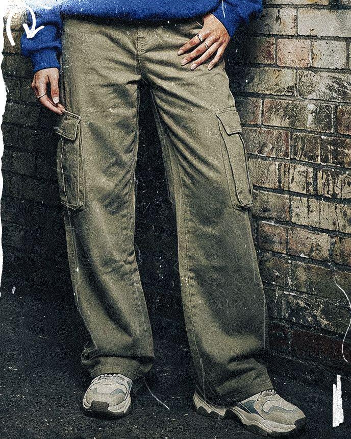 Person wearing green cargo pants and gray sneakers stands against a brick wall, with a blue sweater visible at the waist.