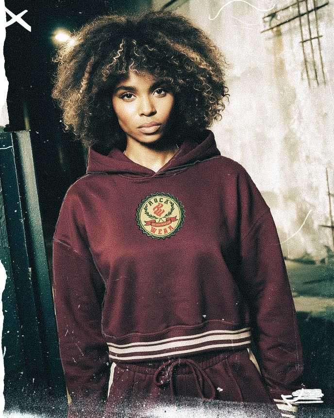 A person with curly hair wears a maroon hoodie with a logo, standing against a textured, urban wall background.