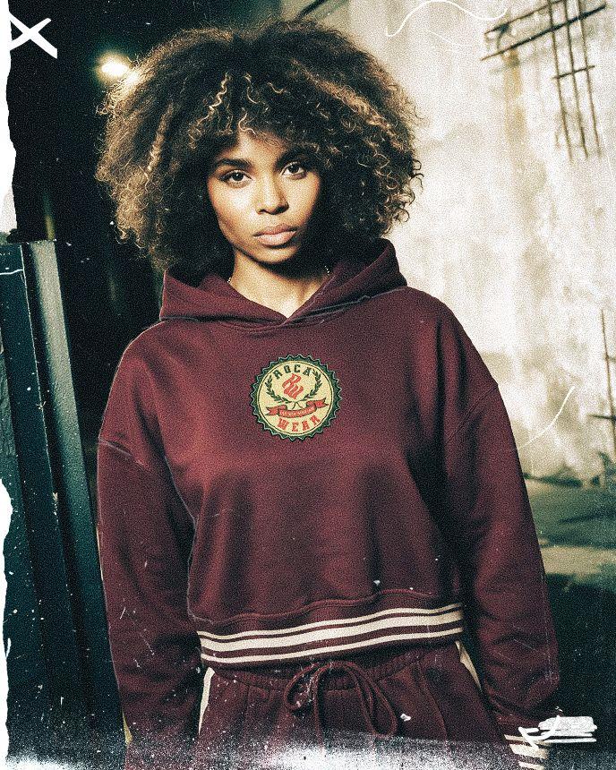 A person with curly hair wears a maroon hoodie with a logo, standing against a textured, urban wall background.
