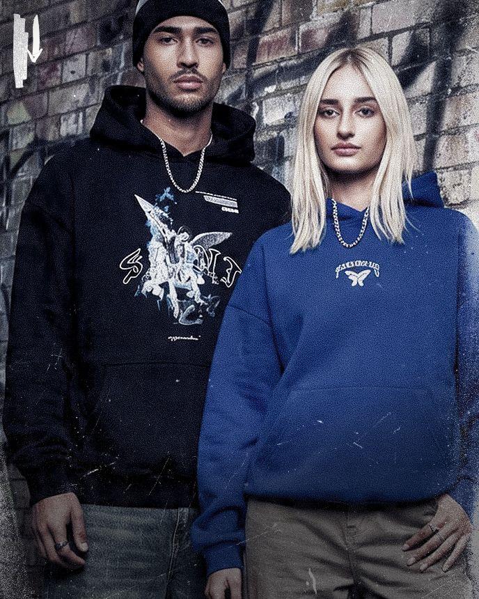 Two people in hoodies, one black and one blue, stand against a graffiti-covered brick wall, wearing silver chains.