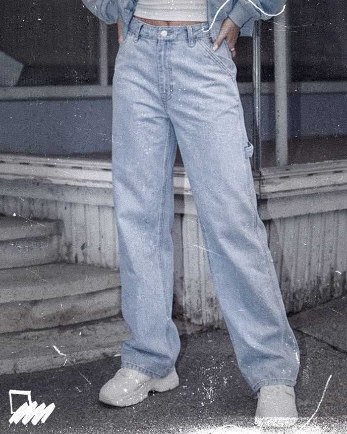 Person in light blue baggy jeans and white sneakers, hands in pockets, standing outside on a worn sidewalk.