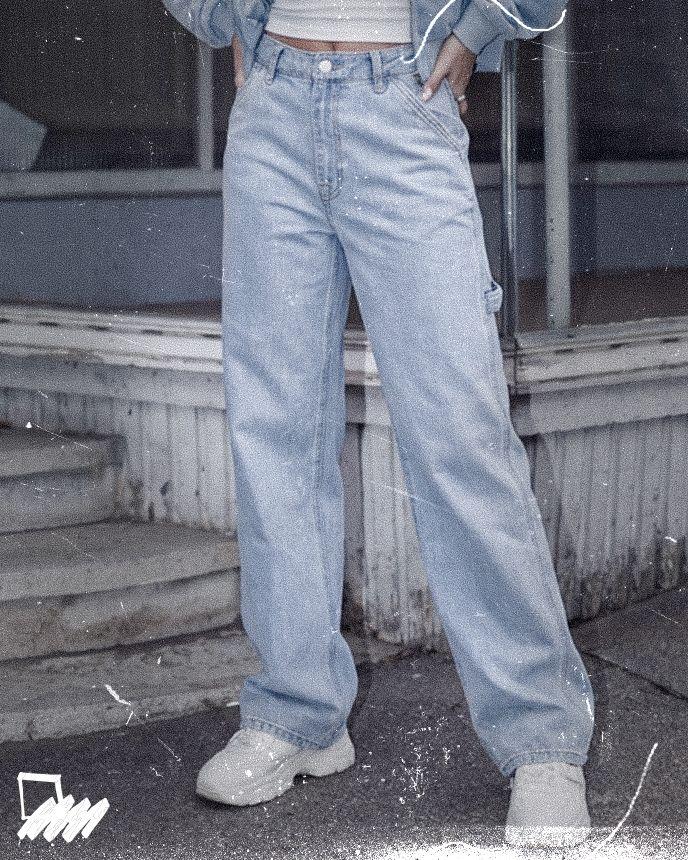 Person in light blue baggy jeans and white sneakers, hands in pockets, standing outside on a worn sidewalk.