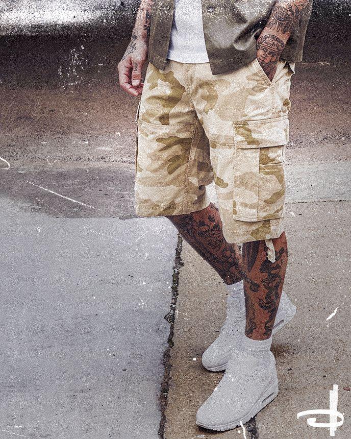 Person wearing camo cargo shorts, white sneakers, and a jacket. Arms and legs have tattoos. Worn texture overlays the image.