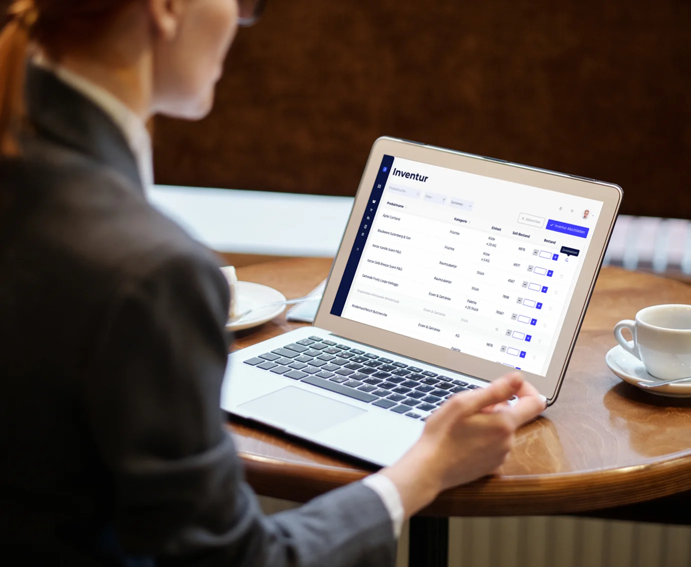 Picture of a woman looking at a grey laptop that's laying on a brown table between several cups. On the laptop screen there is a view from the Avisio hotel inventory management application built by Monterail.