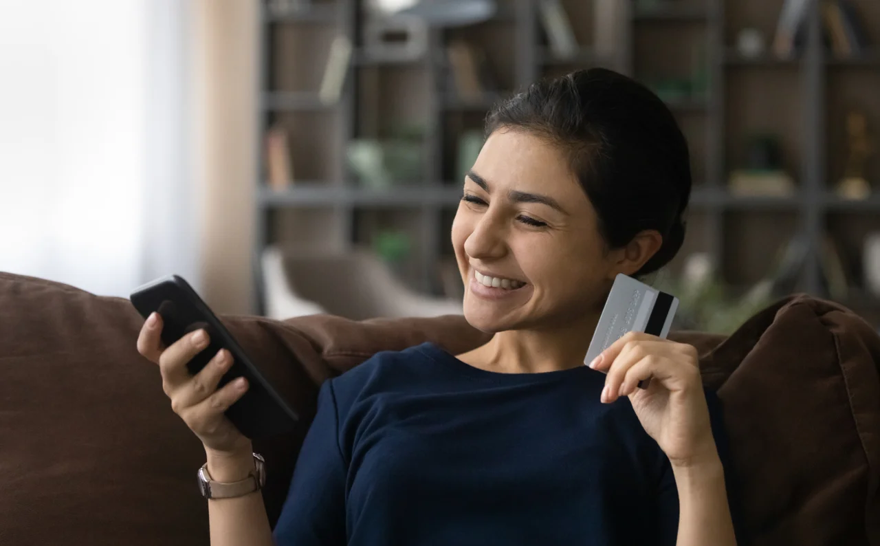 Picture of a woman smiling and holding a phone in one hand and a banking card in the other.