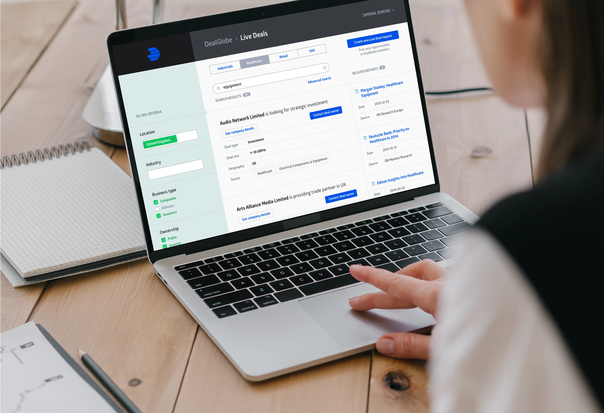 Picture of woman working on a laptop on a wooden desk with several notebooks spread around. On the laptop screen is a view from the DealGlobe application built by Monterail.