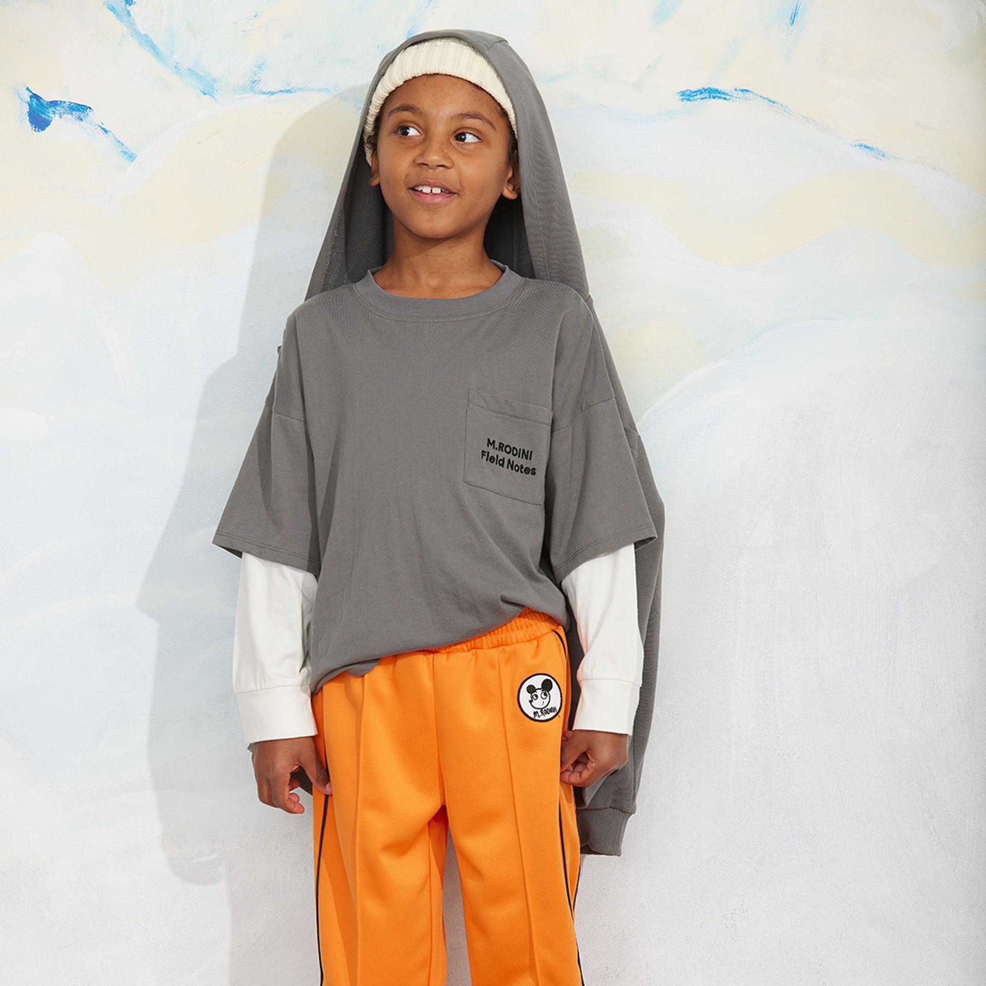 A child wearing a gray T-shirt and orange pants stands against a pastel background, with a gray hoodie draped over their head and a beanie.