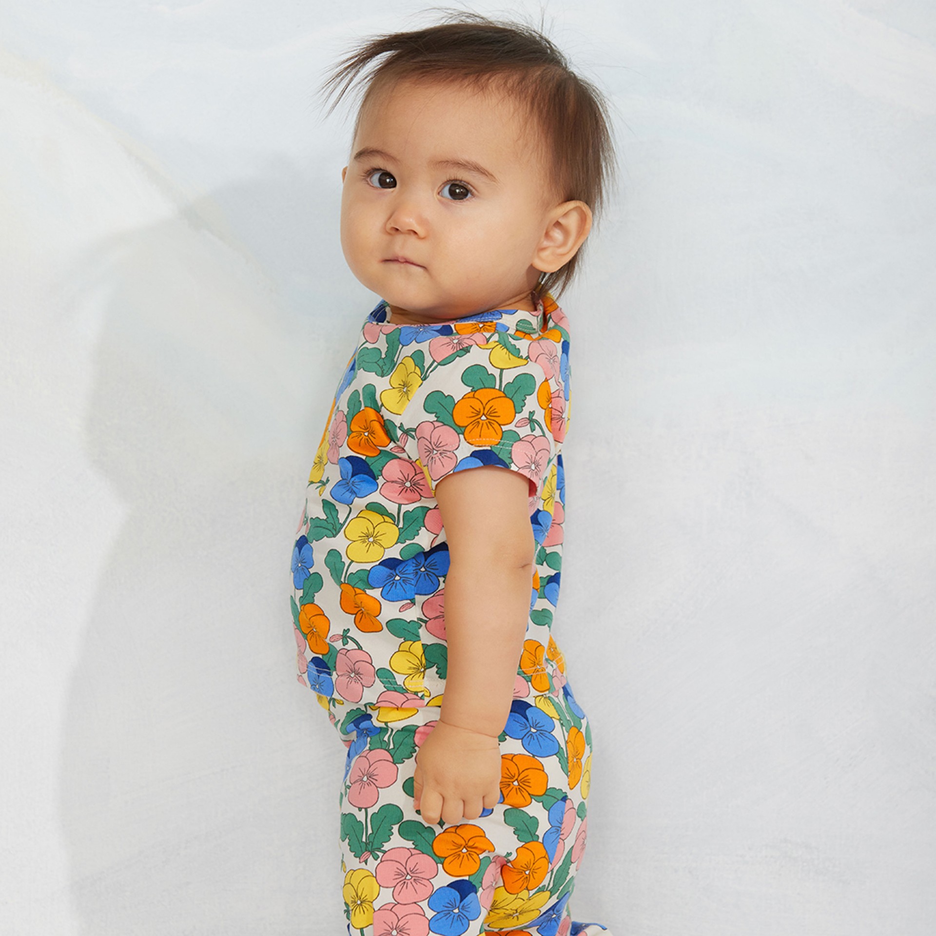 A baby in a floral-patterned outfit stands against a light background, gazing sideways.