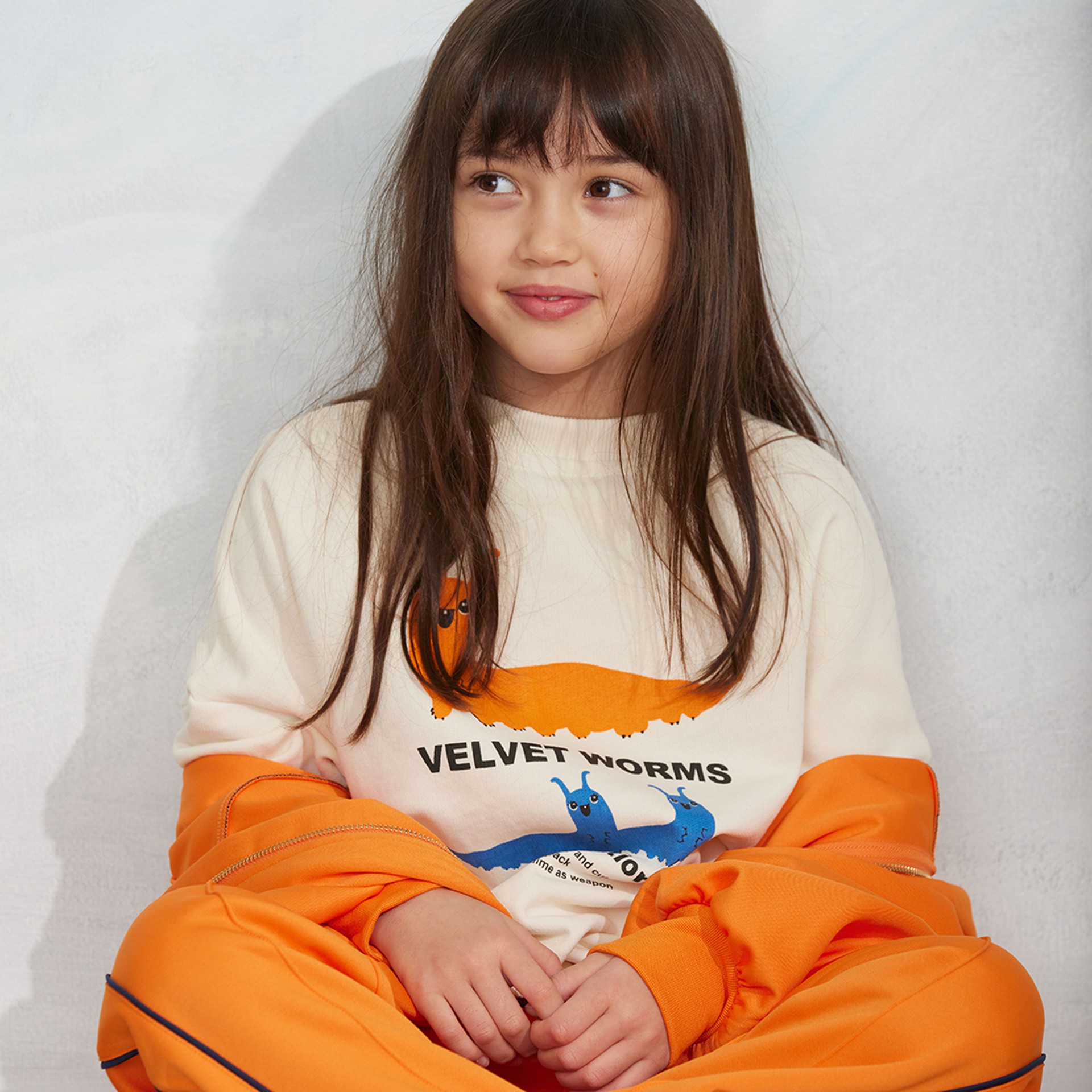 Young girl with long brown hair wearing a cream sweater with orange and blue velvet worm design and orange pants, sitting against a light background.