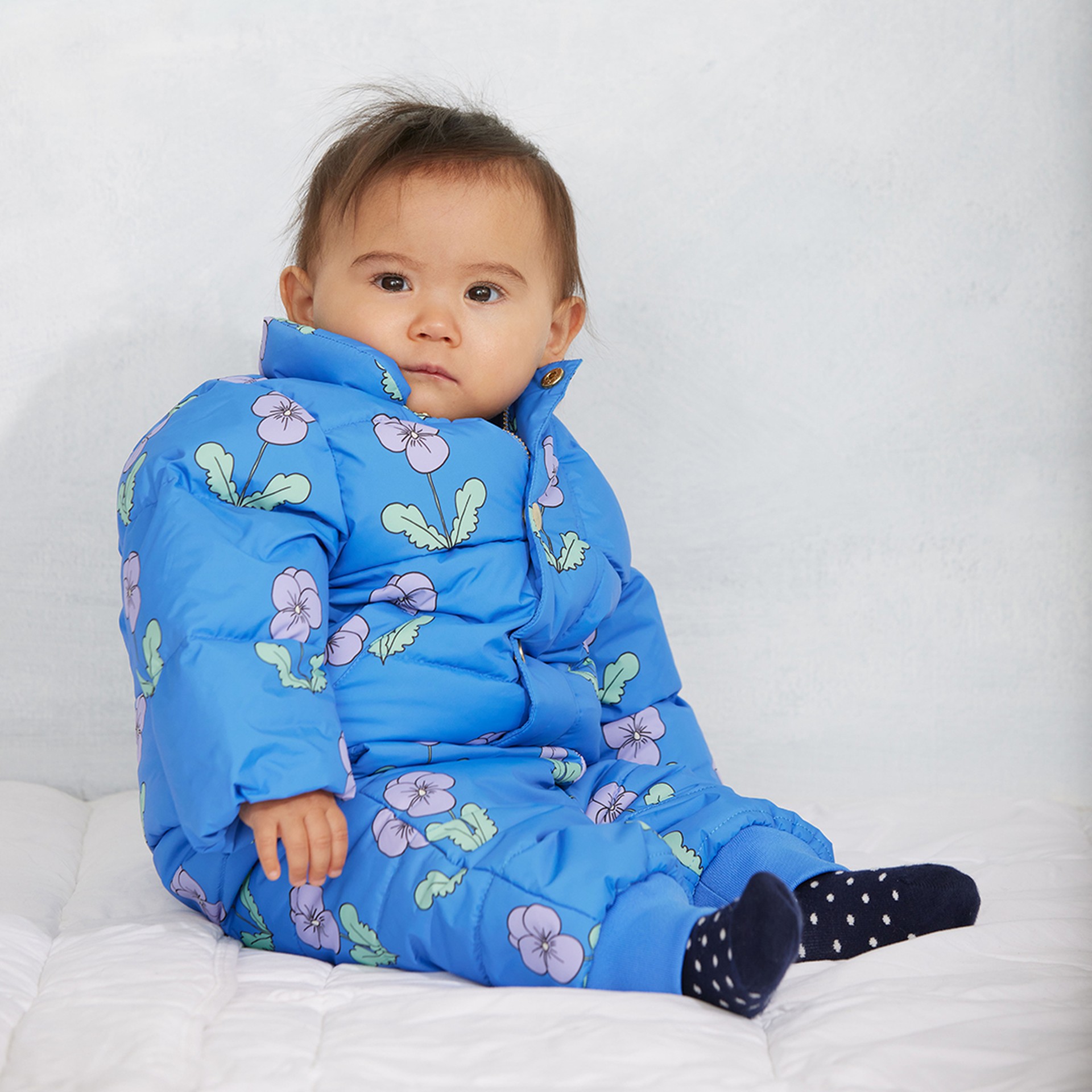 A baby in a blue jacket with floral patterns sits on a white surface, looking at the camera.