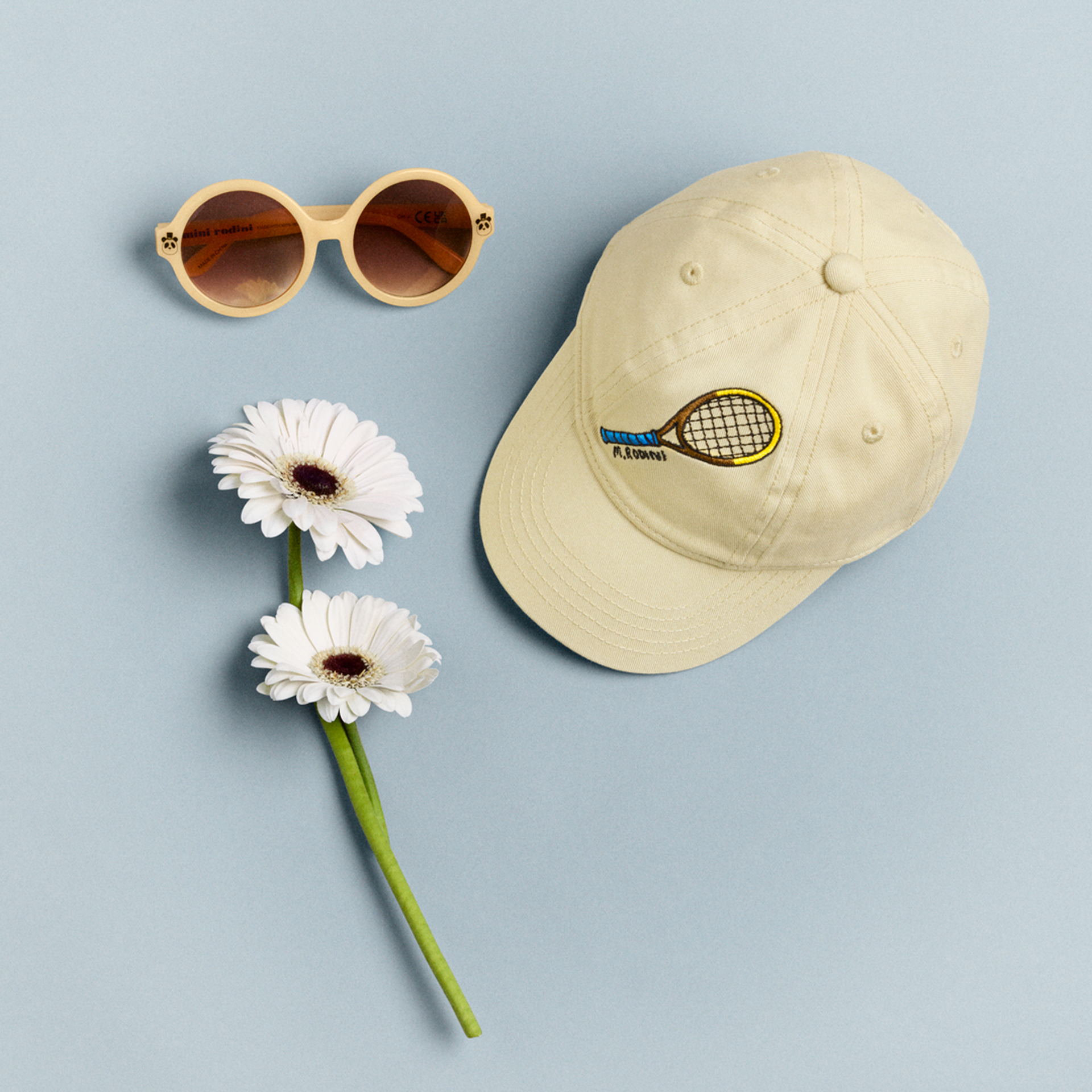 Beige cap with tennis racket design, round sunglasses, and two white daisies on a light blue background.