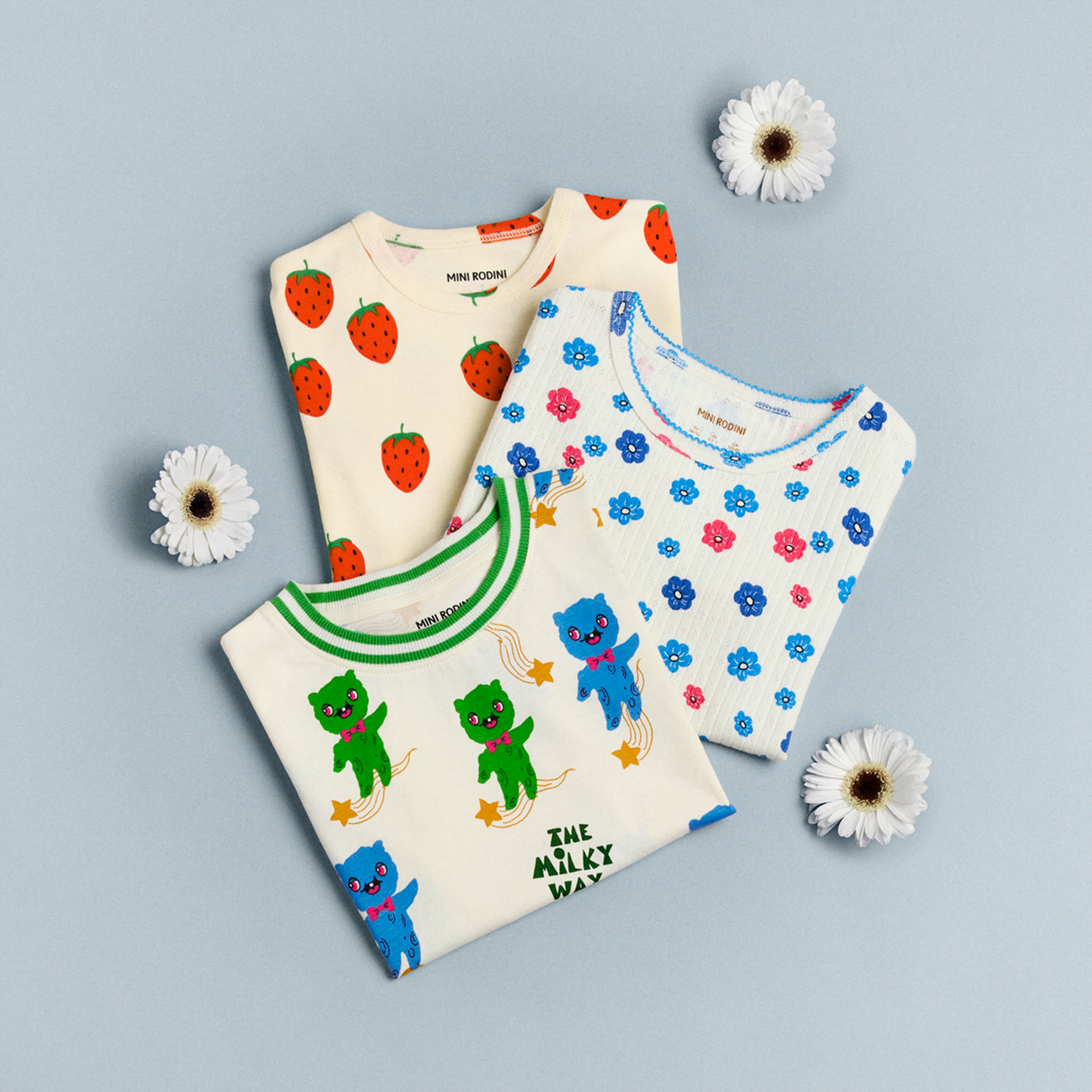 Three colorful children's shirts with strawberry, floral, and playful bear designs are laid out on a light blue background with daisies.