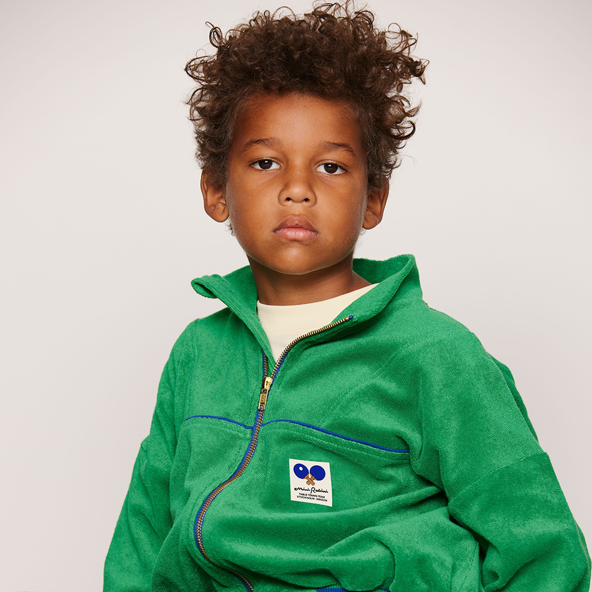 Young child with curly hair wearing a green zip-up jacket from Mini Rodini looking directly at the camera against a neutral background.