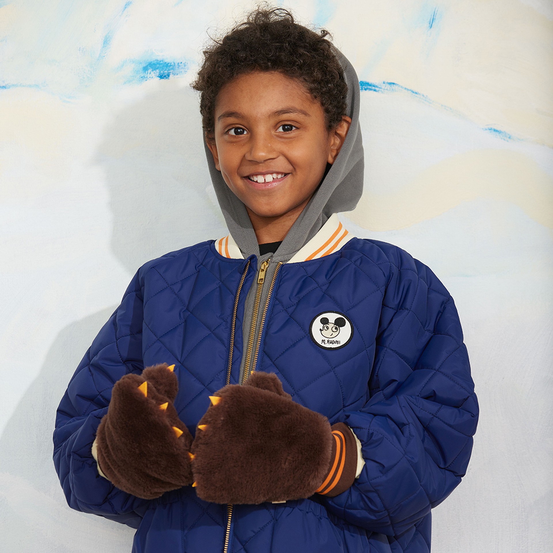 A smiling child in a blue quilted jacket with a hoodie, wearing brown furry mittens resembling bear claws, standing against a light background.