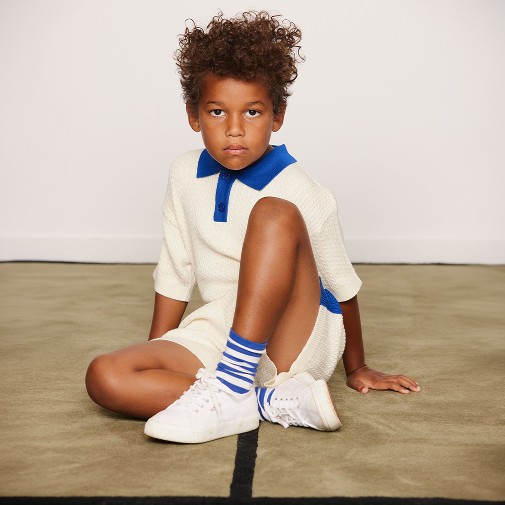 Child sitting on a beige carpet wearing a cream outfit with blue details, blue-striped socks all from Mini Rodini looking at the camera.