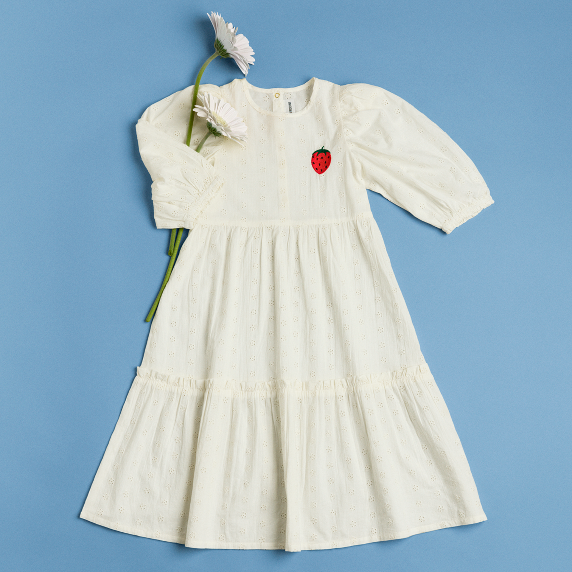 White eyelet dress with puff sleeves and strawberry patch on the chest, accompanied by white daisies, on a blue background.