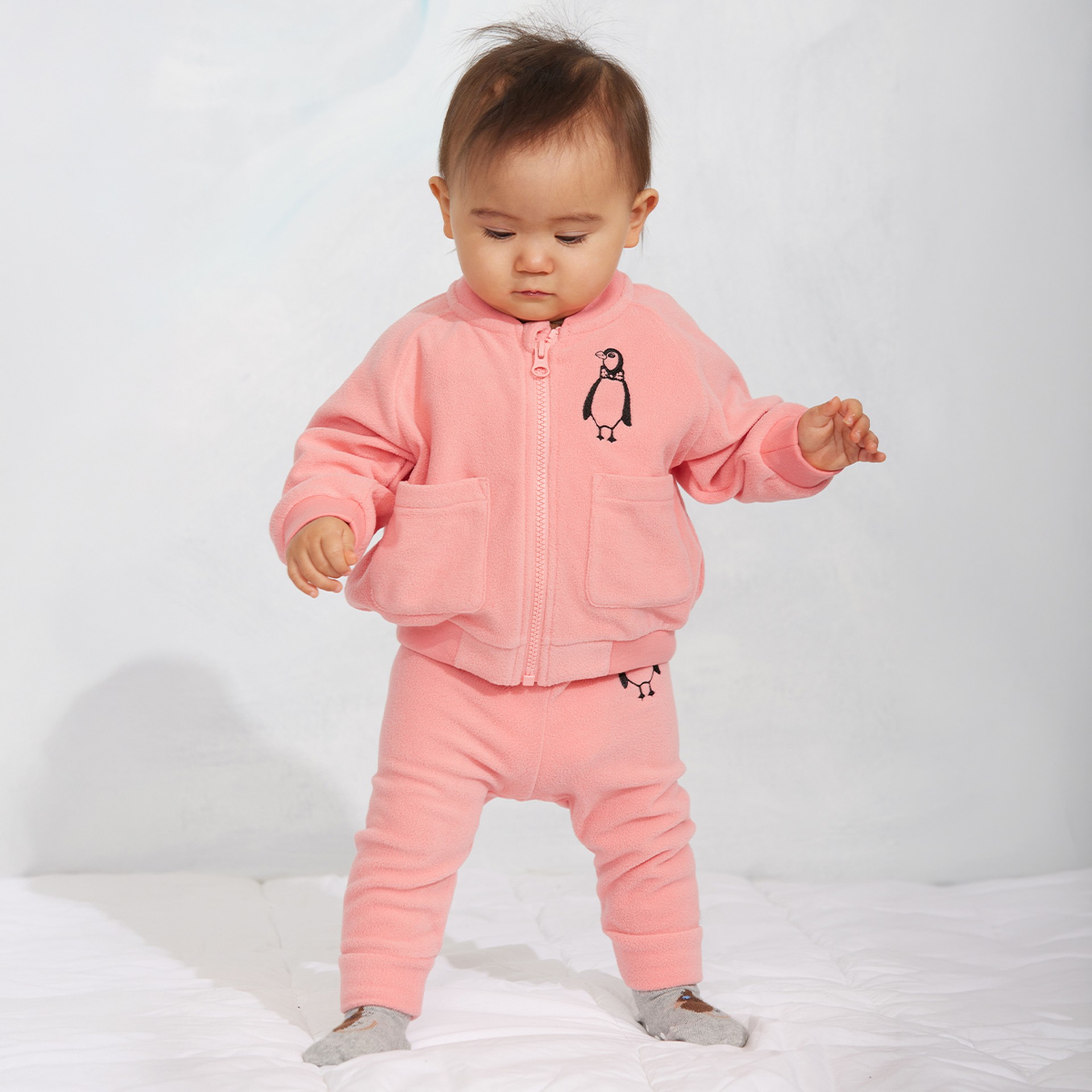 A toddler wearing a pink fleece outfit with penguin designs stands on a white surface, looking down with one hand slightly raised.
