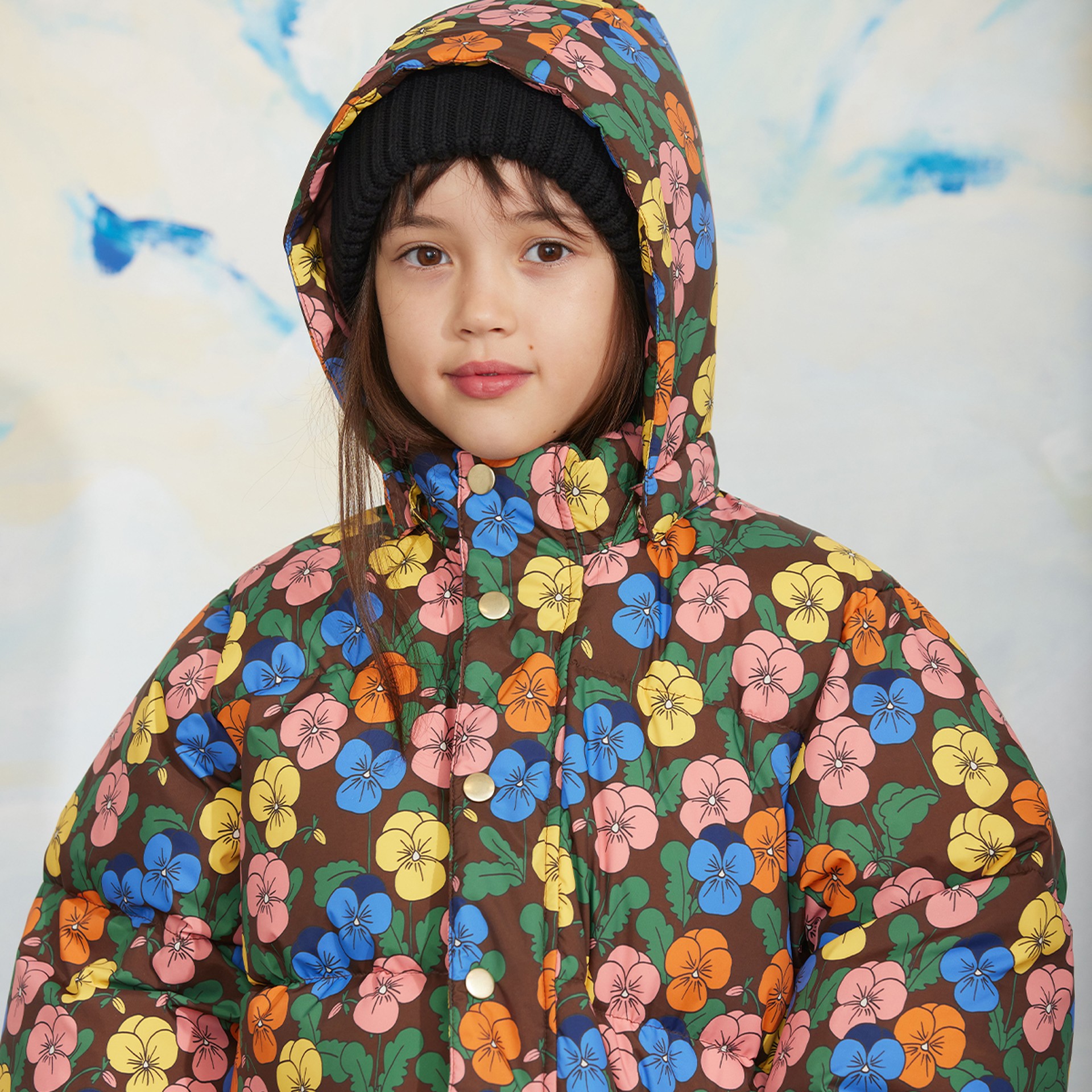 Child wearing a colorful floral-patterned hooded jacket and a black knit hat, standing against a light background.