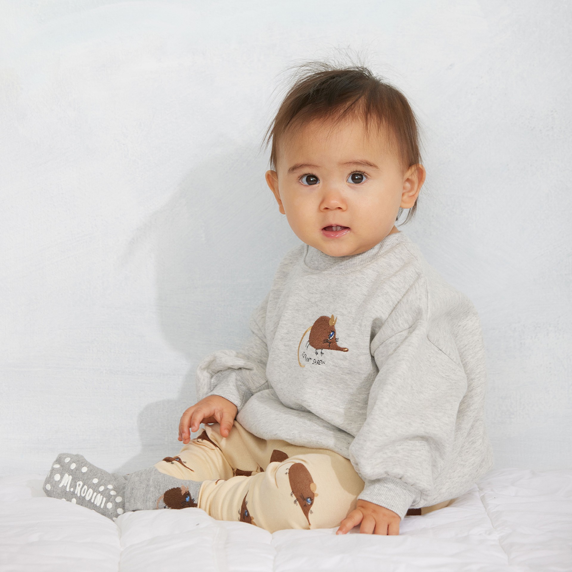 A baby in gray and beige clothing with bear designs sits on a white quilt, looking at the camera.