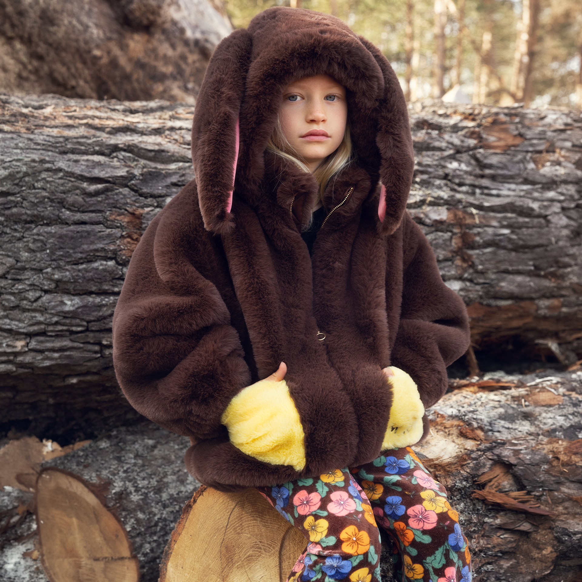 A child sits on a log, wearing a furry brown coat with bunny ears and yellow mittens, paired with colorful floral pants in a forest setting.