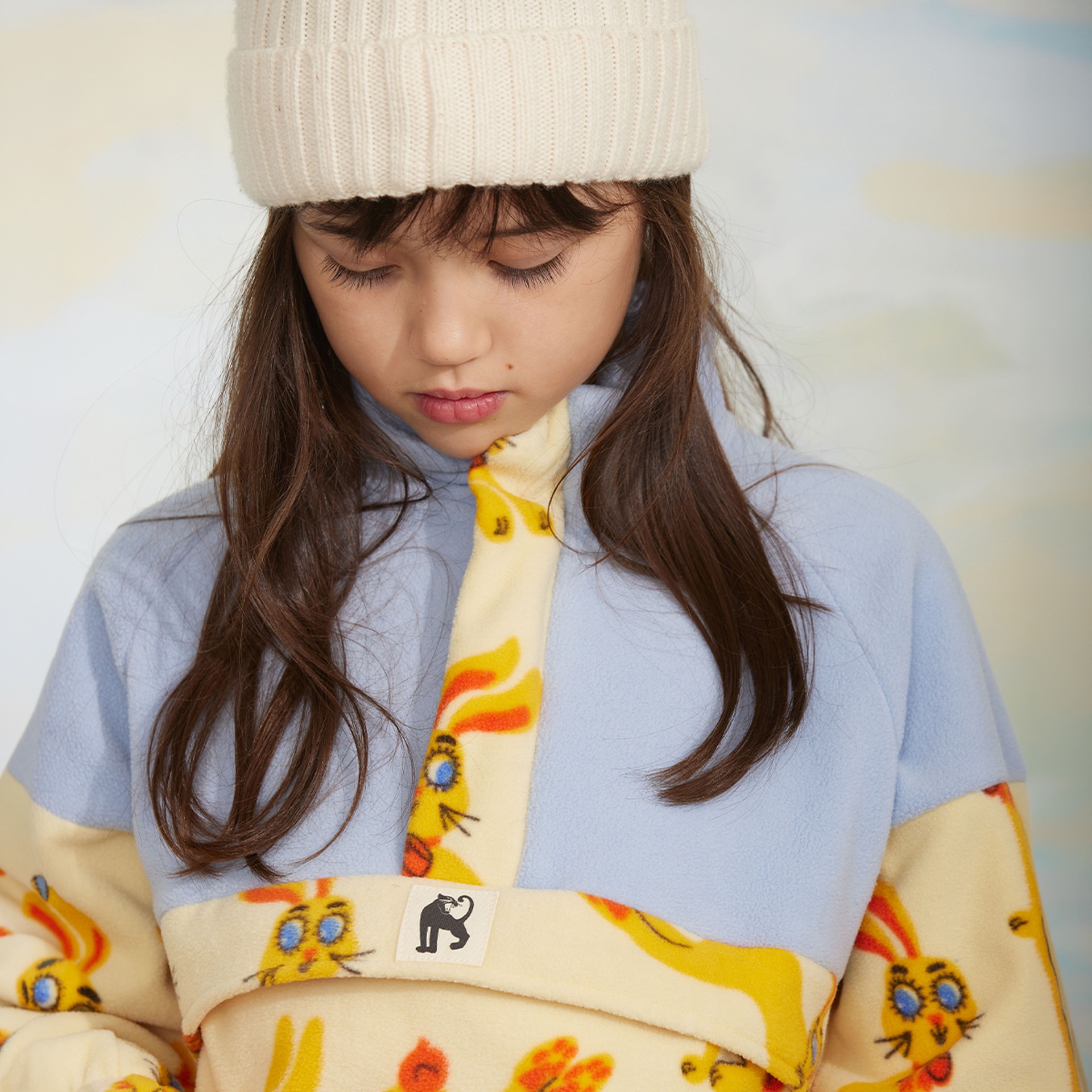 A young girl with long brown hair wearing a cream knit hat and a fleece jacket with colorful rabbit designs looks down.