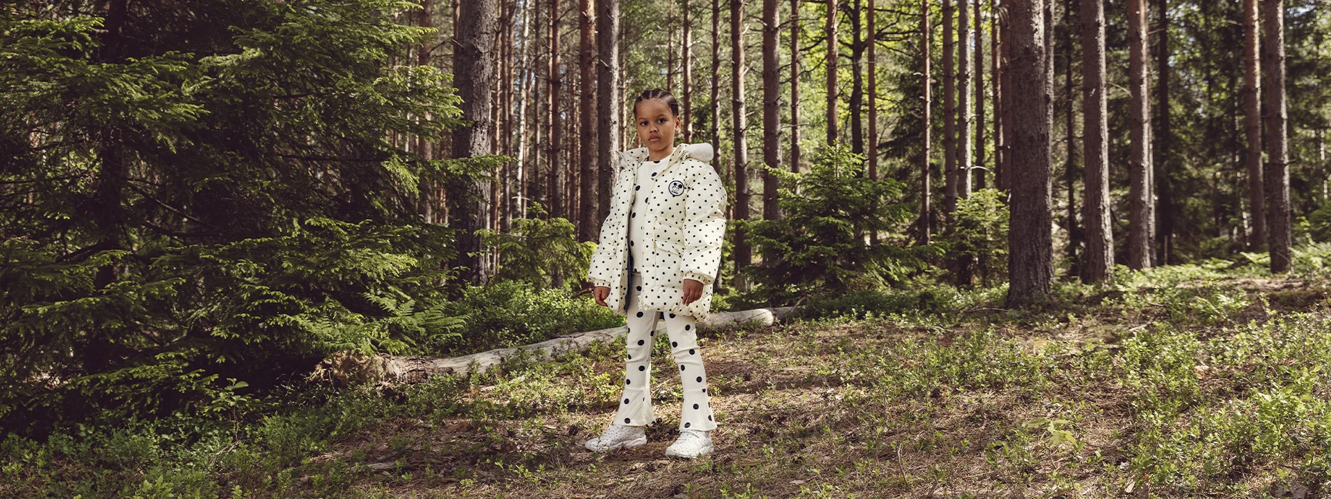 A child stands in a forest wearing a white polka dot outfit, including a jacket and pants, surrounded by trees and greenery.