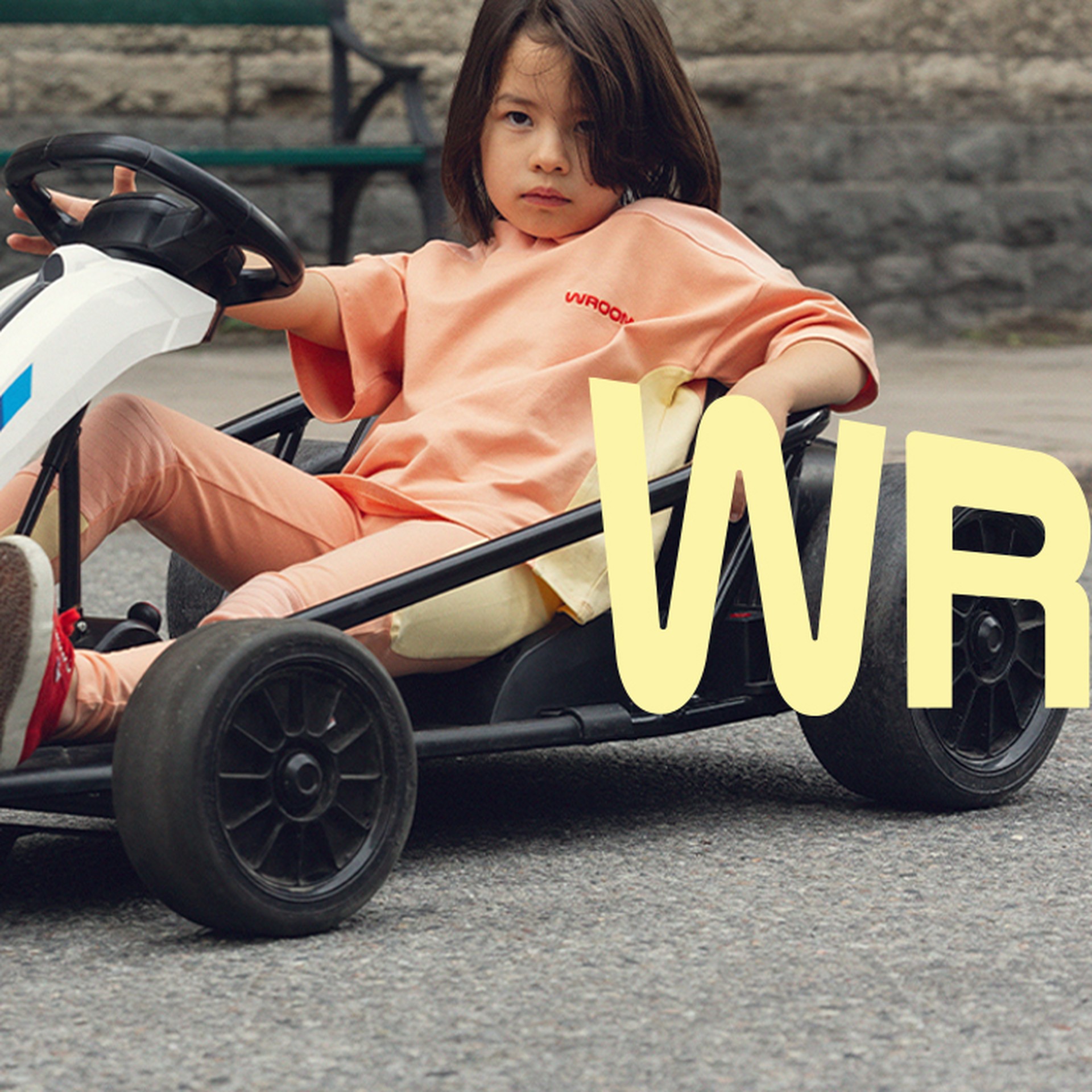 Child sitting in a small white go-kart on a street, wearing an orange outfit. The word "WROOM" overlays the image.