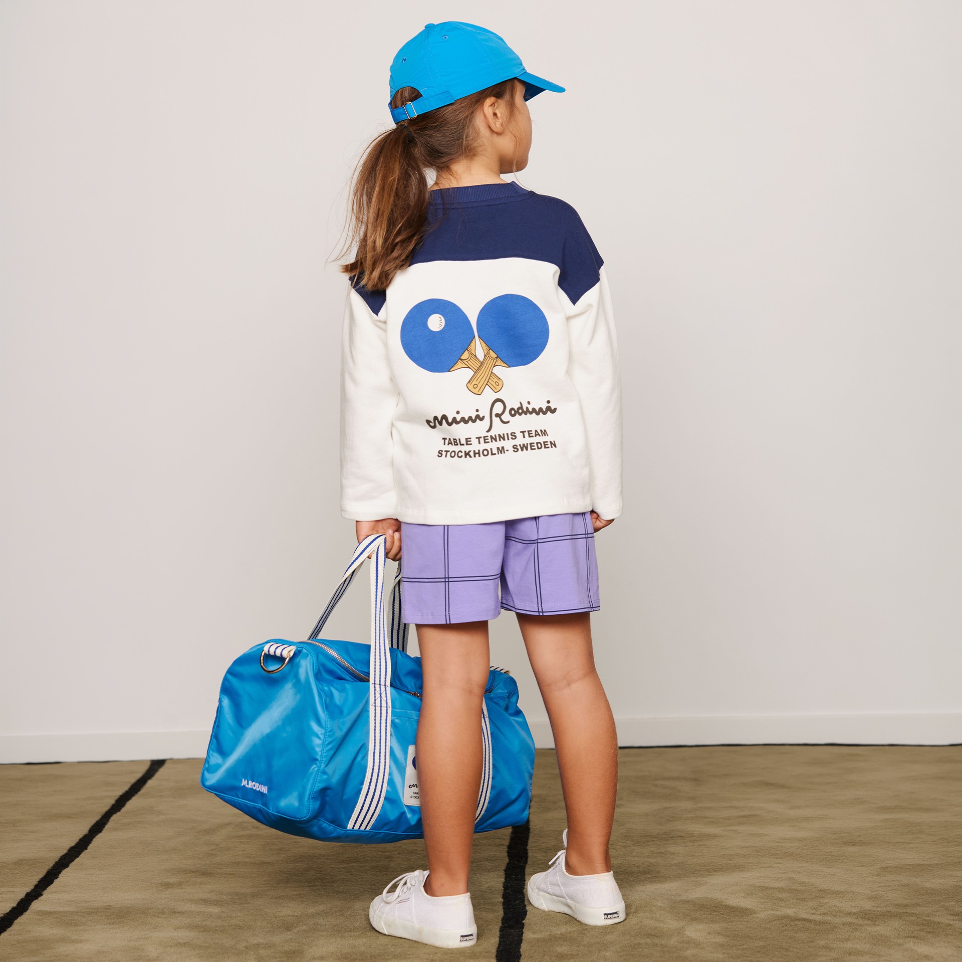 Child in Table tennis patch nylon cap, Table tennis gym bag and purple shorts, facing away, holding a blue duffel bag. Sweater has table tennis graphic and text about a team from Stockholm. Outfit from Mini Rodini.