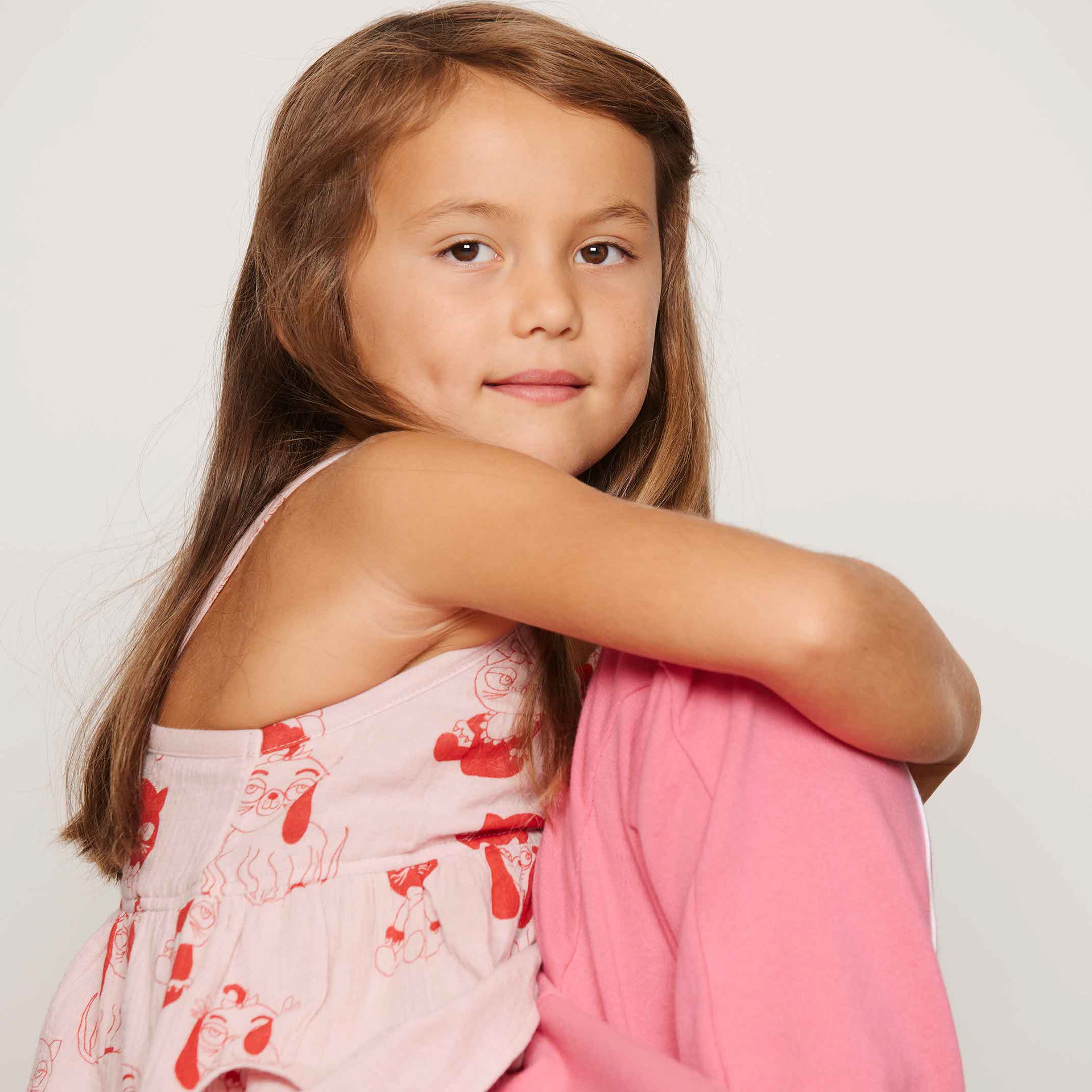 Young girl with long brown hair, wearing a pink top with Mini Babies print and plain piping sweatpants from Mini Rodini, gazes softly at the camera against a plain background.