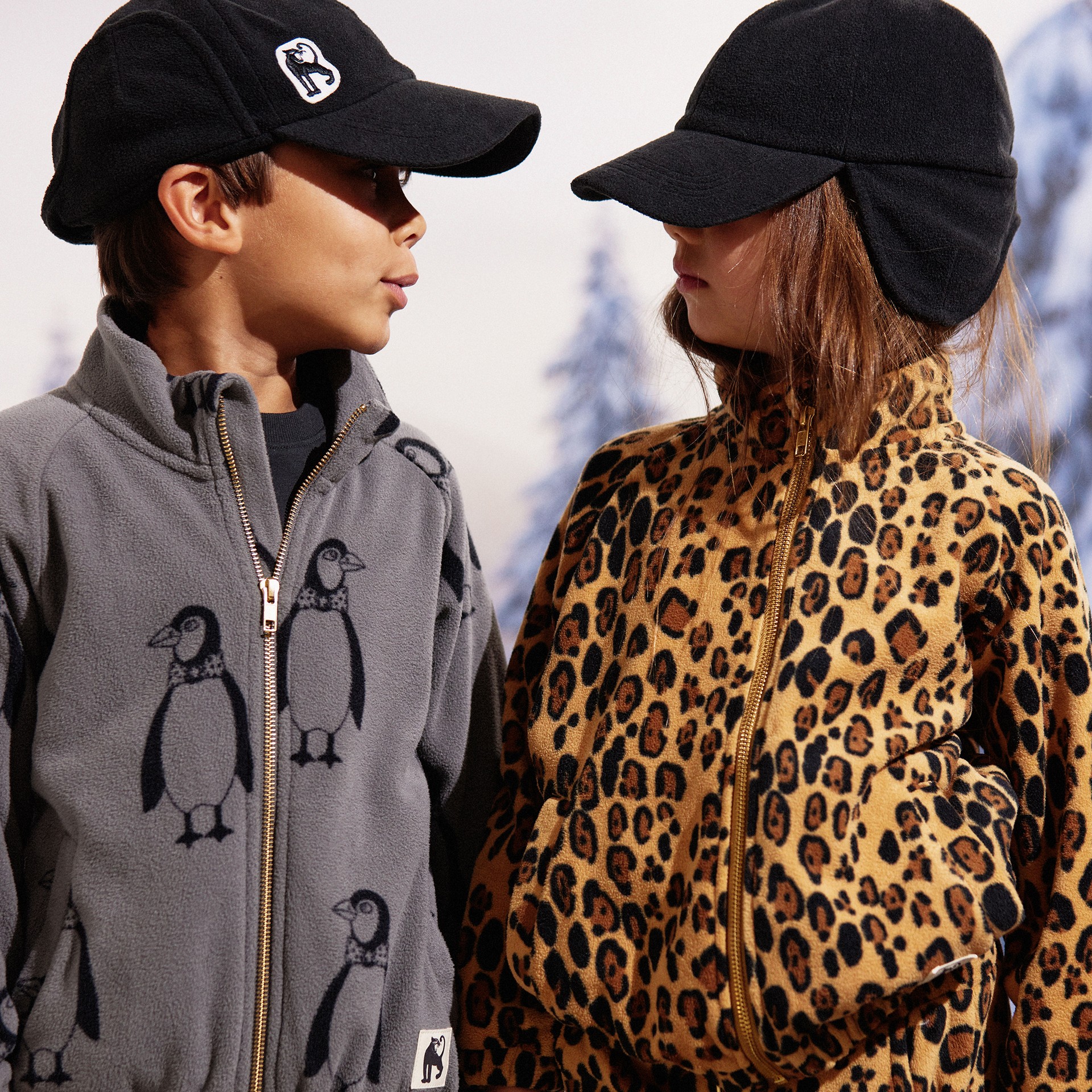 Two children wearing black caps and fleece outfits, one in grey with penguin prints and the other in leopard print, facing each other.