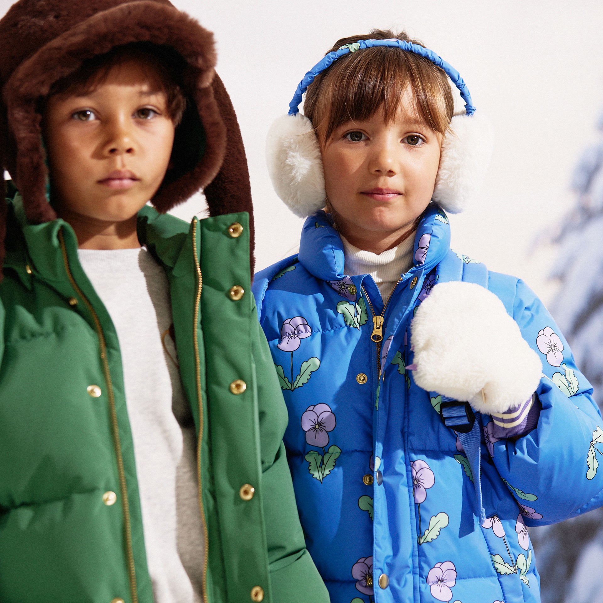 Two children in winter clothing; one wears a green coat and brown hood, the other wears a blue floral coat with earmuffs and gloves.