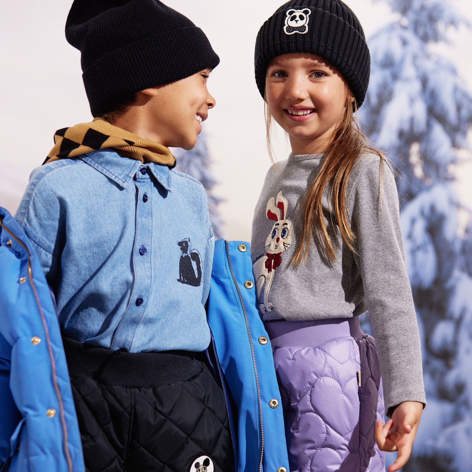 Two children in winter clothing, wearing black beanies, smiling at each other with a snowy backdrop. One wears a blue coat, the other a gray sweater.