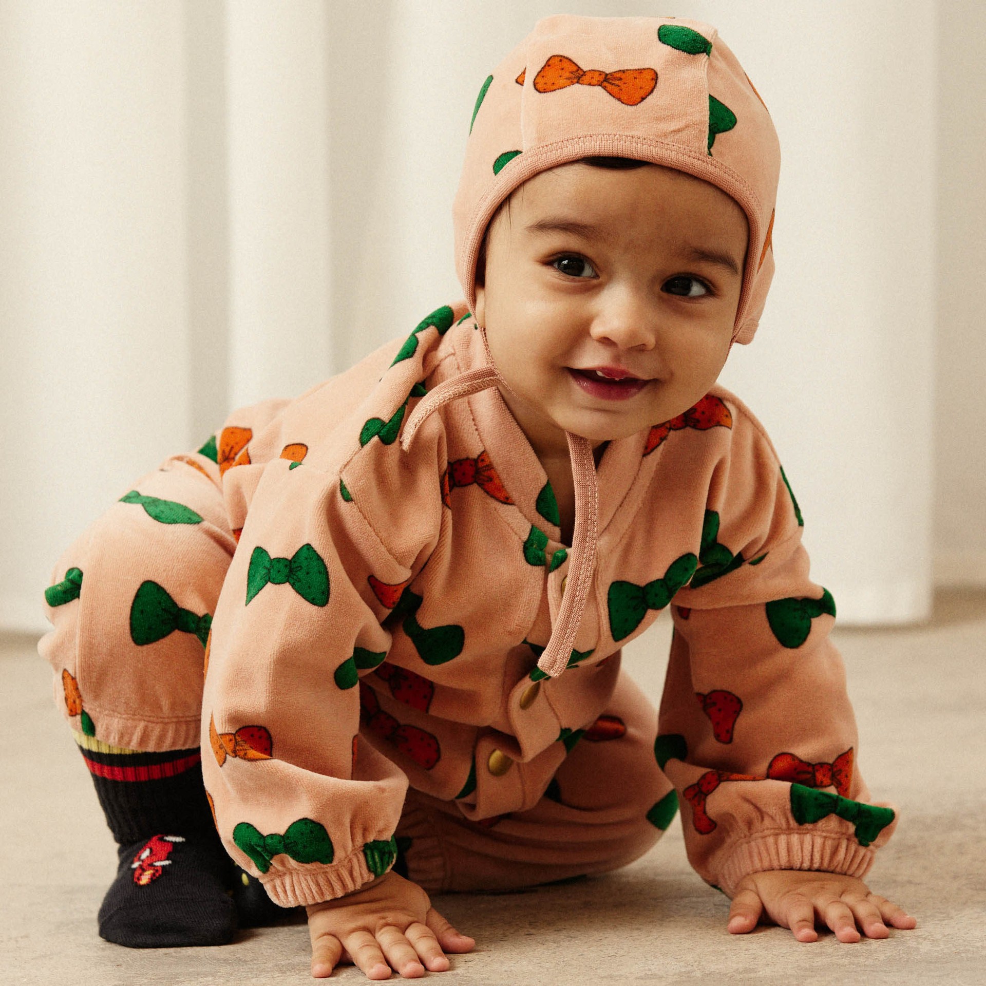 A baby in a pink outfit with colorful bow tie patterns, crawling on the floor, smiling, and wearing a matching hat and socks.