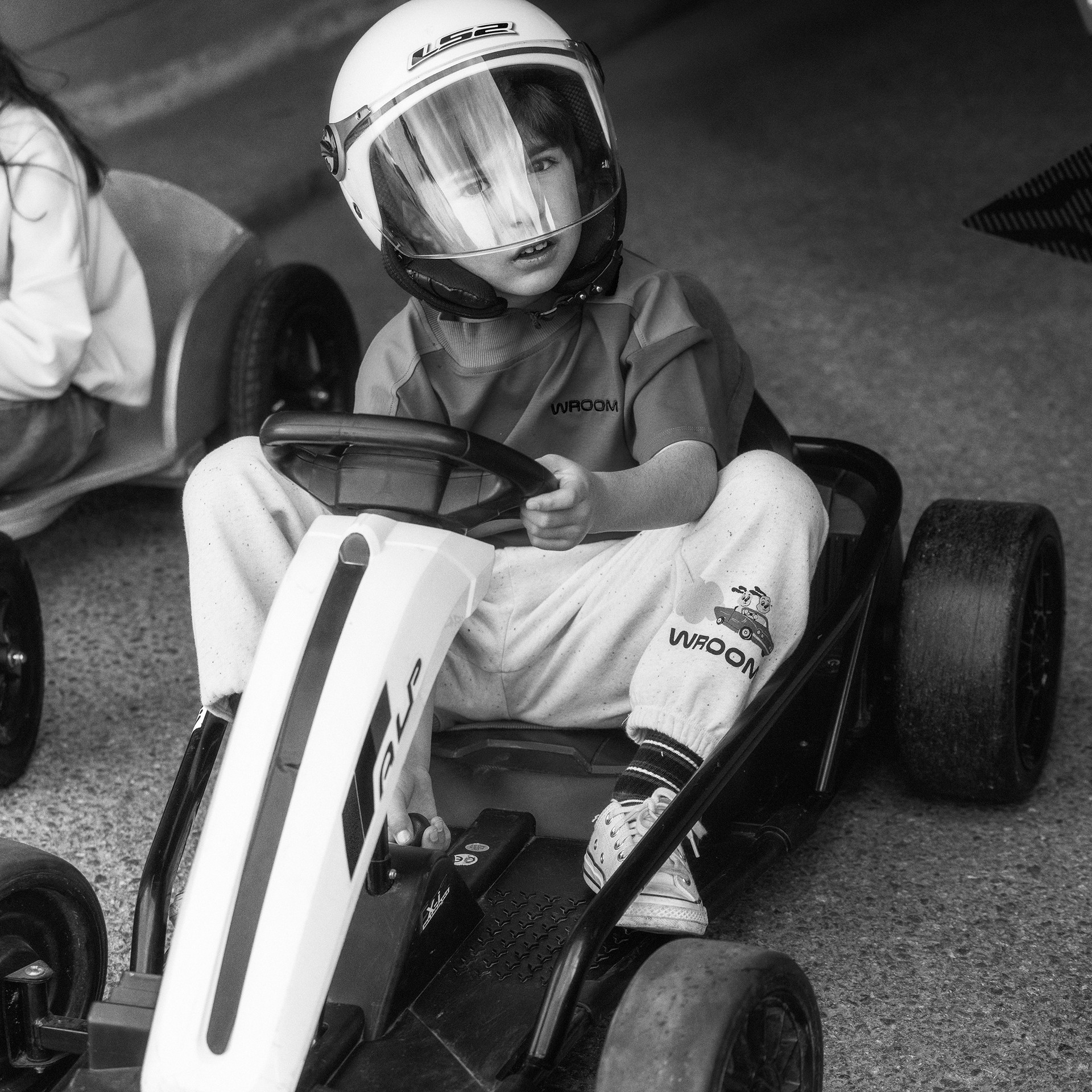 A child wearing a helmet drives a small go-kart, looking focused. The monochrome image highlights the kart and the child's determined expression.