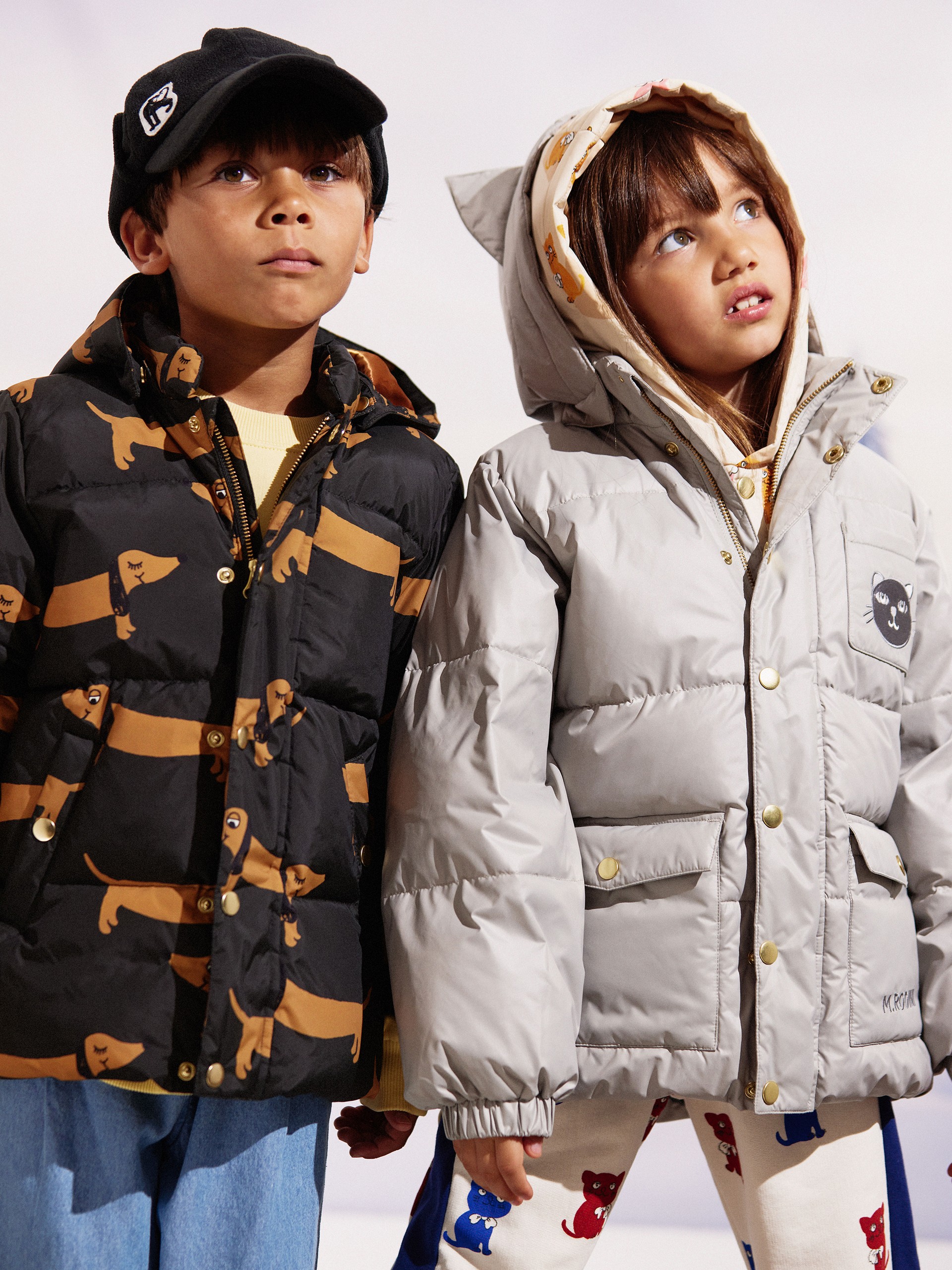 Two children in winter jackets, one with a dachshund print and the other in gray with cat-ears, stand closely together looking upward.