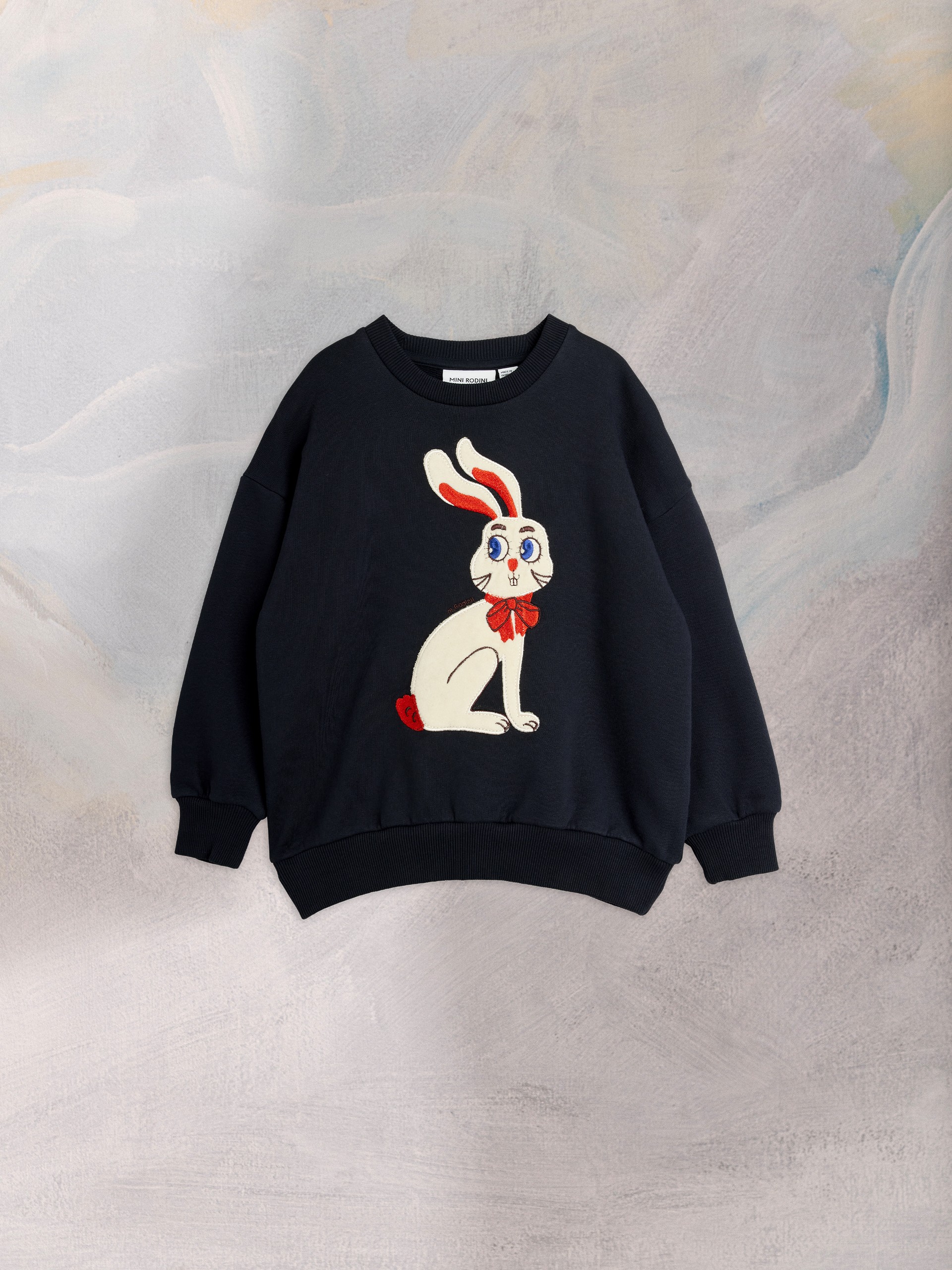Navy blue sweatshirt with a cartoon rabbit design, featuring red ears and a bow tie, set against a textured pastel background.