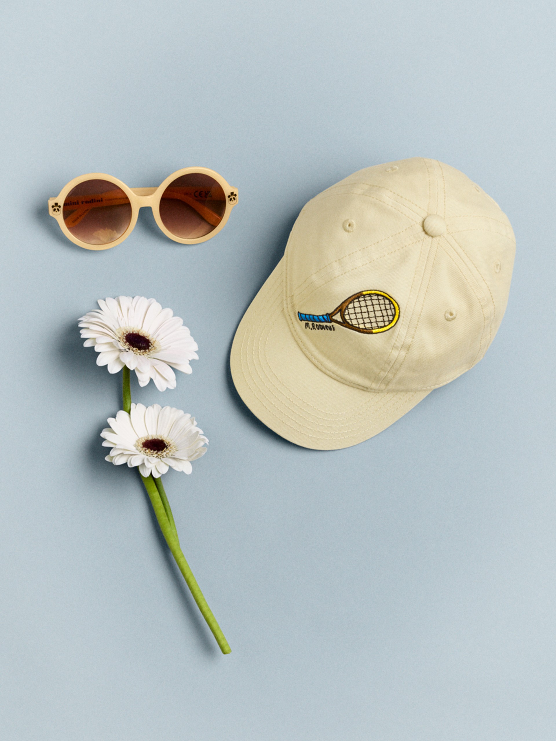 Round sunglasses, a beige cap with a tennis racket embroidery, and two white daisies are arranged on a light blue background.