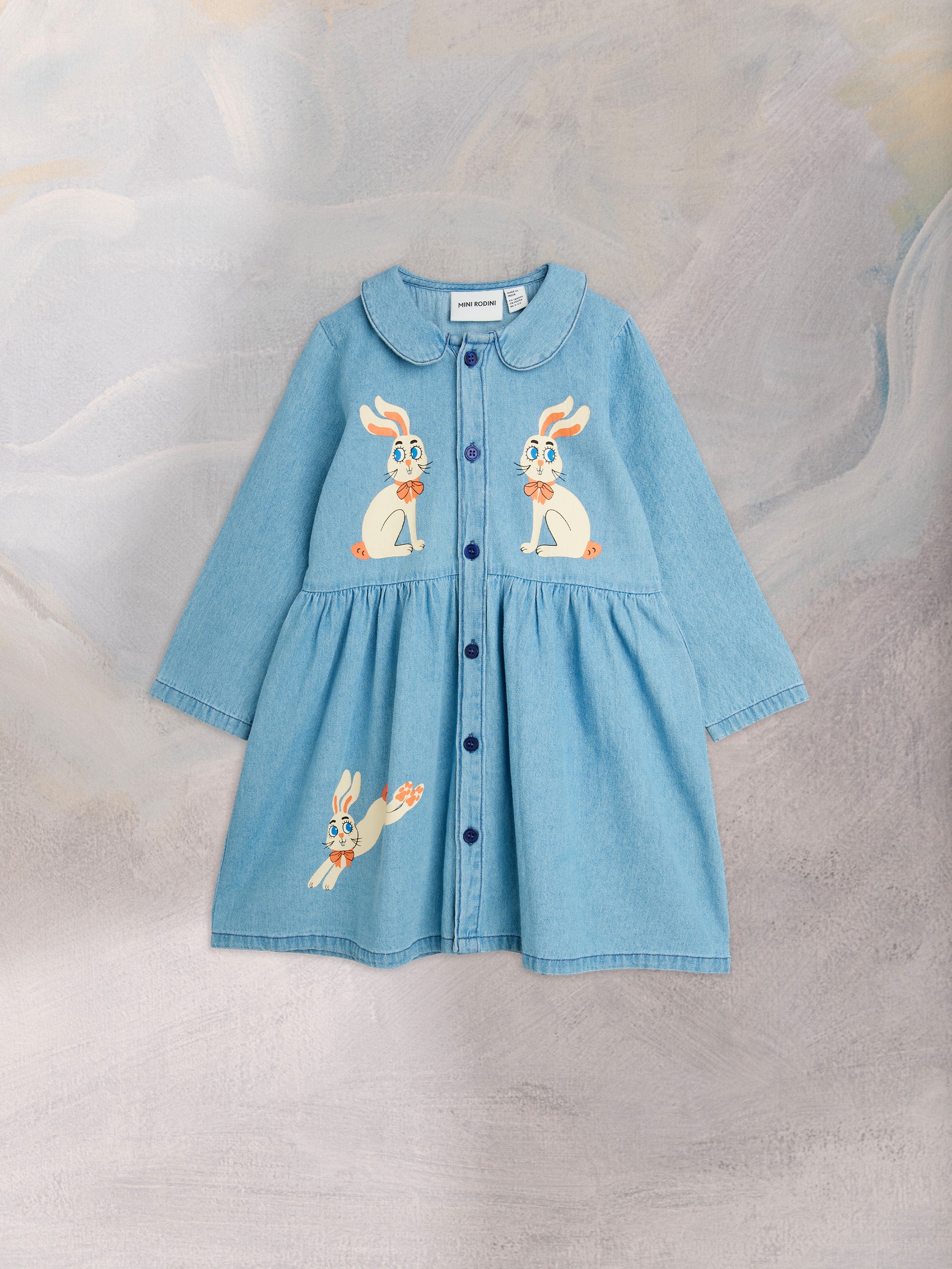 A light blue denim dress with rabbit illustrations, featuring a Peter Pan collar and blue buttons, displayed against a textured backdrop.