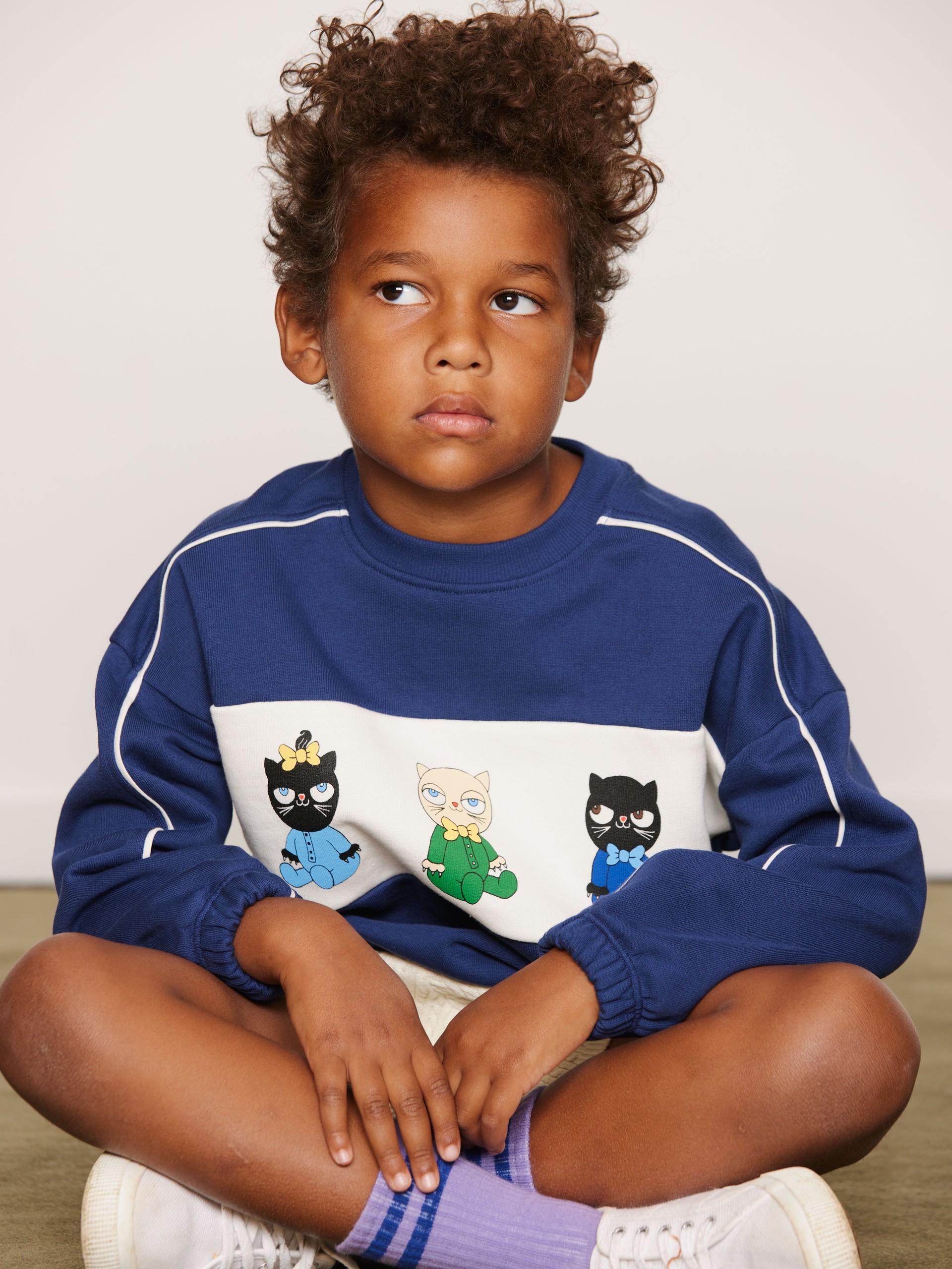 Child in a blue sweatshirt with Minibabies from Mini Rodini, sitting cross-legged on a beige floor, looking to the side.