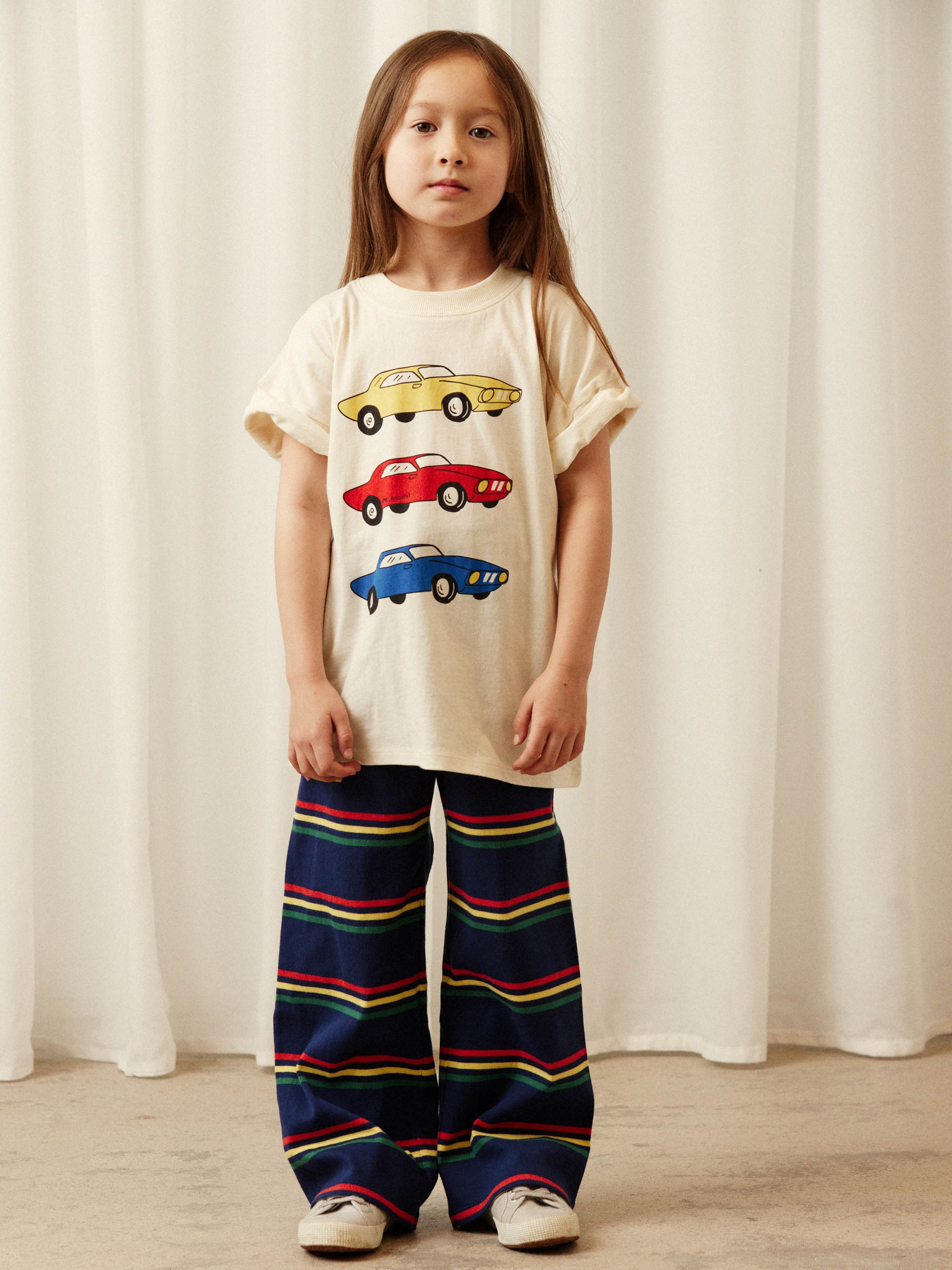 A young child stands indoors wearing a t-shirt with colorful cars and striped wide-legged pants, set against a curtain backdrop.
