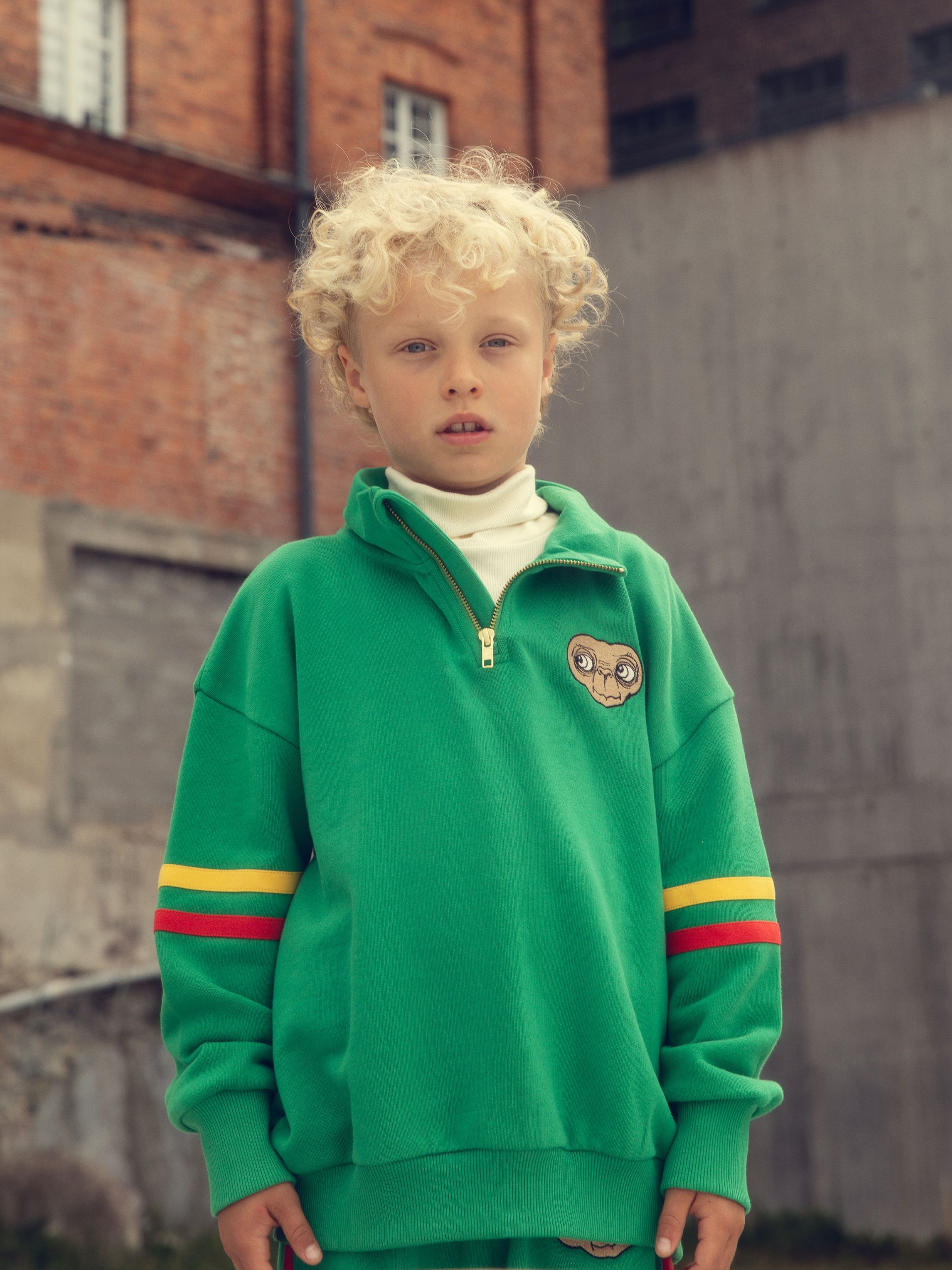 Child with curly hair in a green sweater with stripes and an E.T patch, stands in an urban setting with brick walls in the background. Set from Mini Rodini