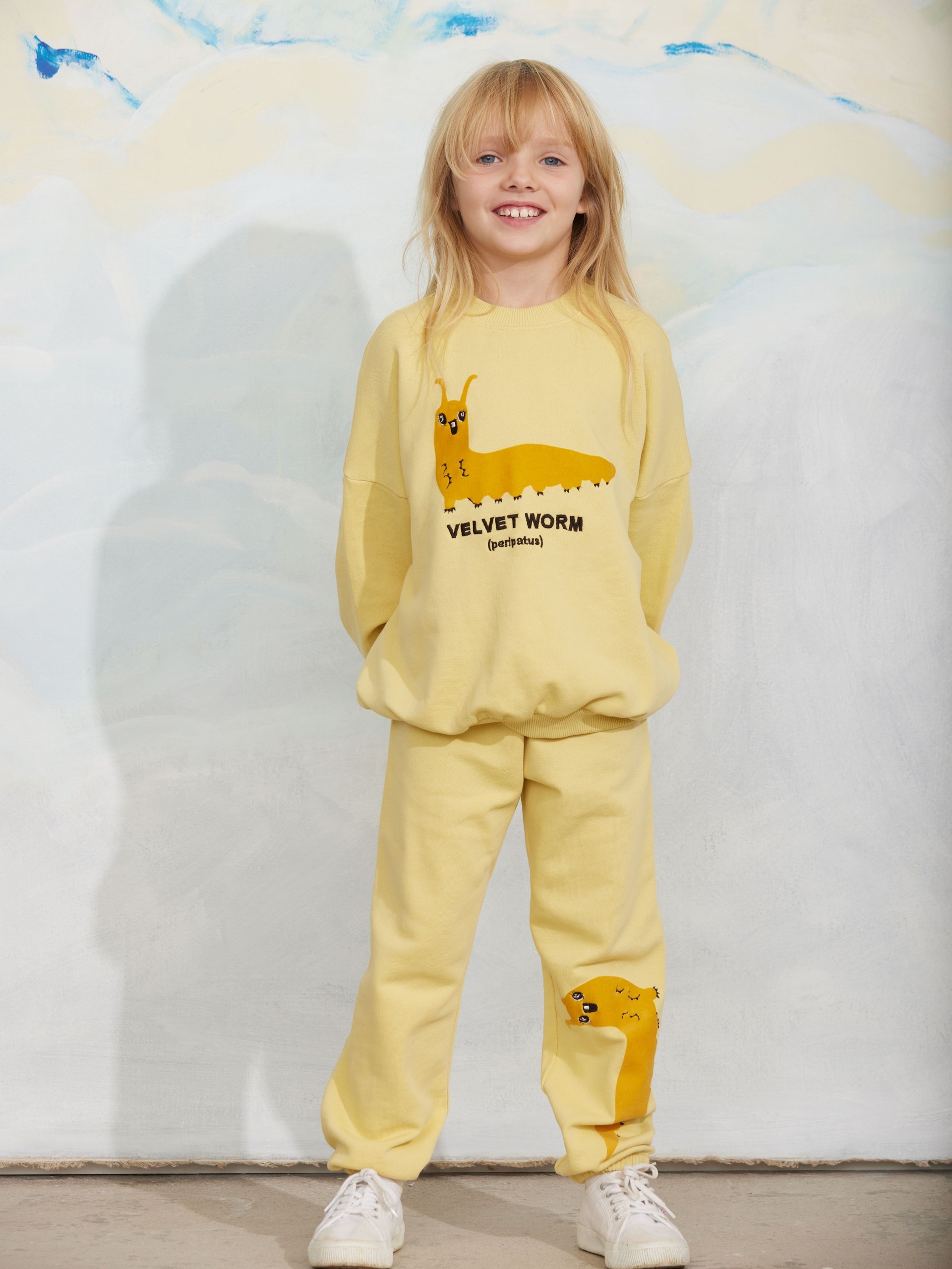 Child smiling in a yellow tracksuit featuring a cartoon velvet worm design, standing against a light painted background.
