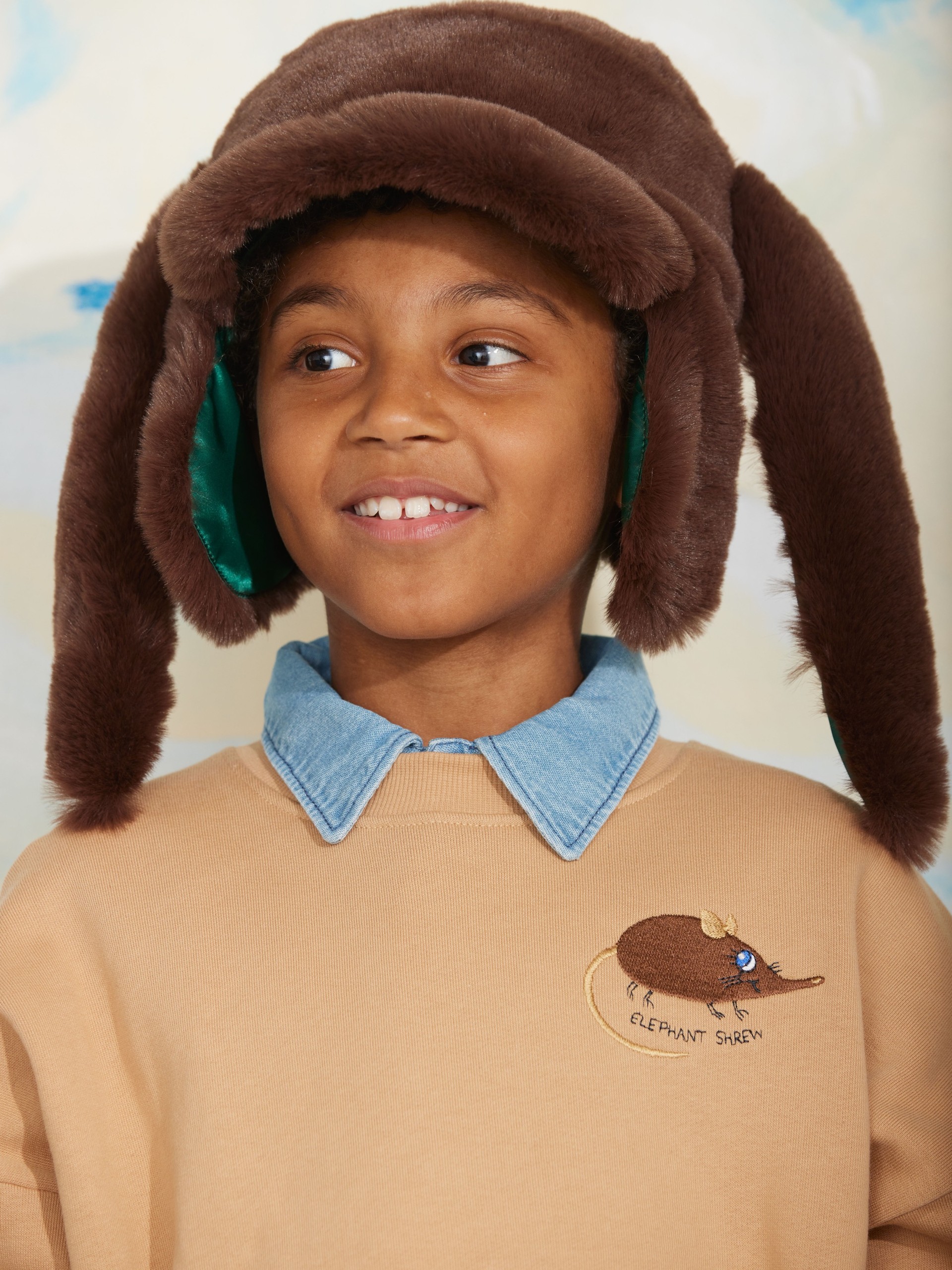 Child wearing a brown furry hat with ear flaps and a tan sweater with an "Elephant Shrew" design. Denim collar peeks out from the sweater.