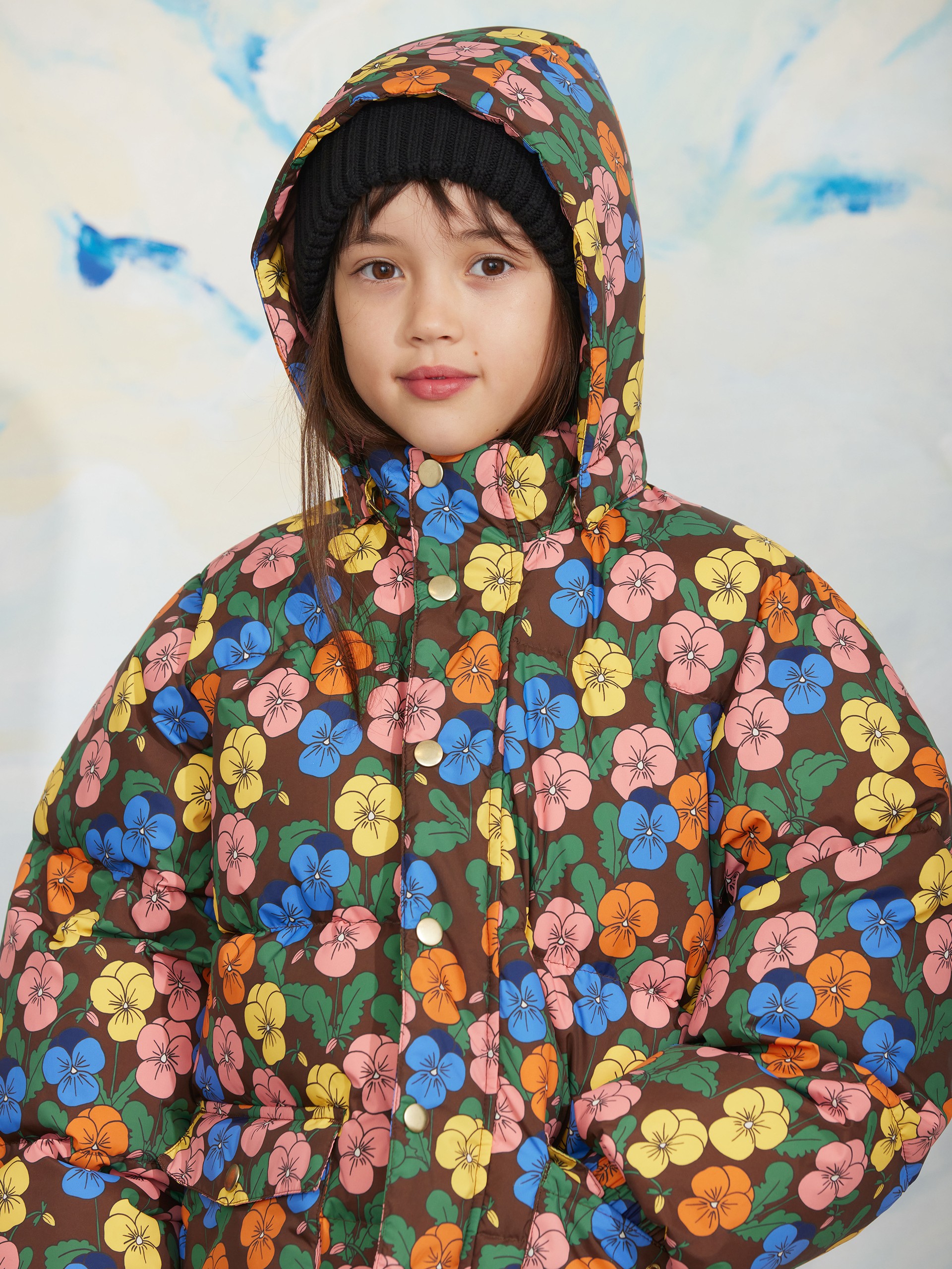 A child wearing a colorful floral-patterned winter jacket and a black knit beanie, standing against a snowy, sky-themed background.