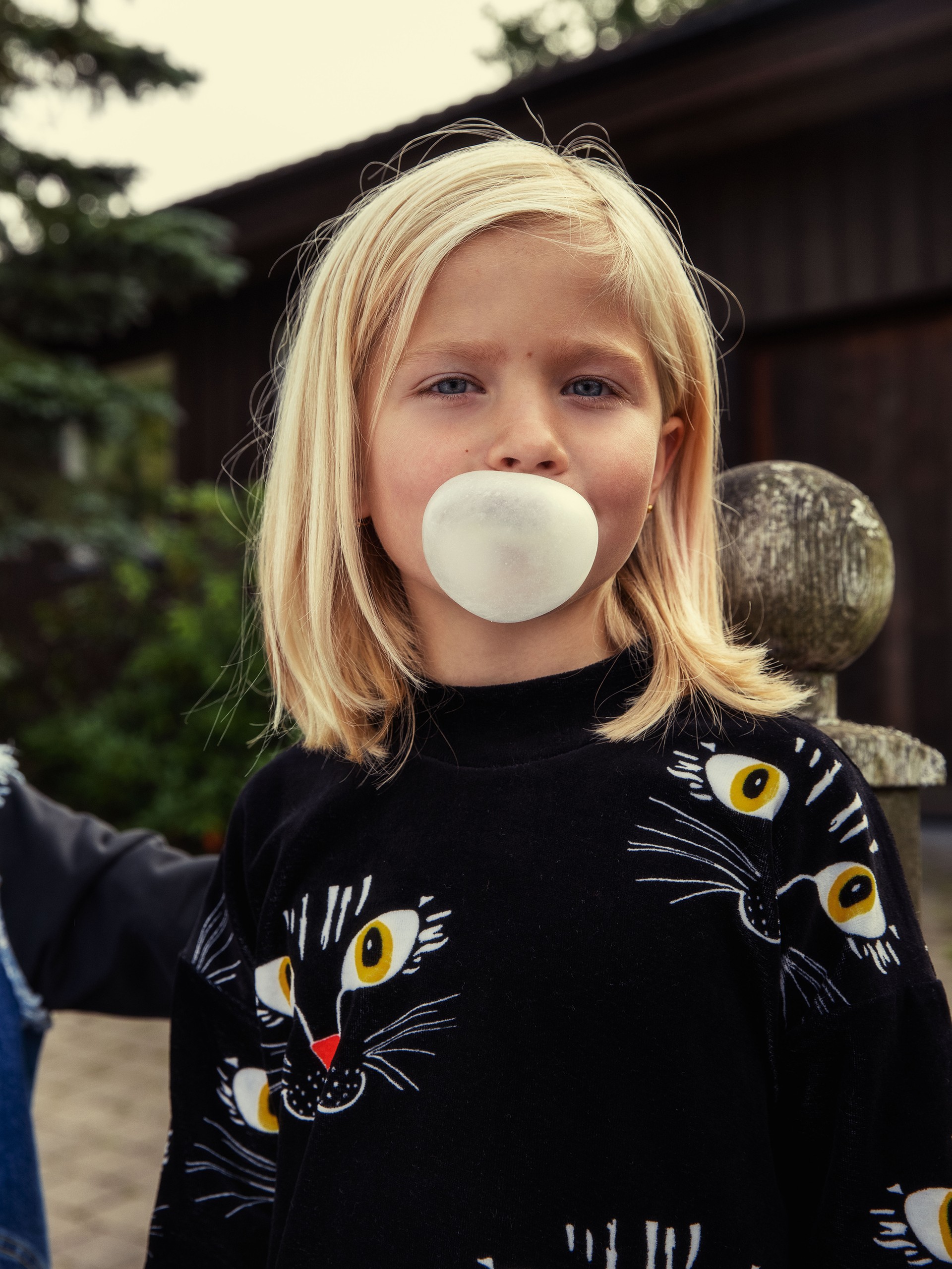 A young child with blonde hair blows a bubble with gum, wearing a black sweater featuring a cat face design, standing outdoors.