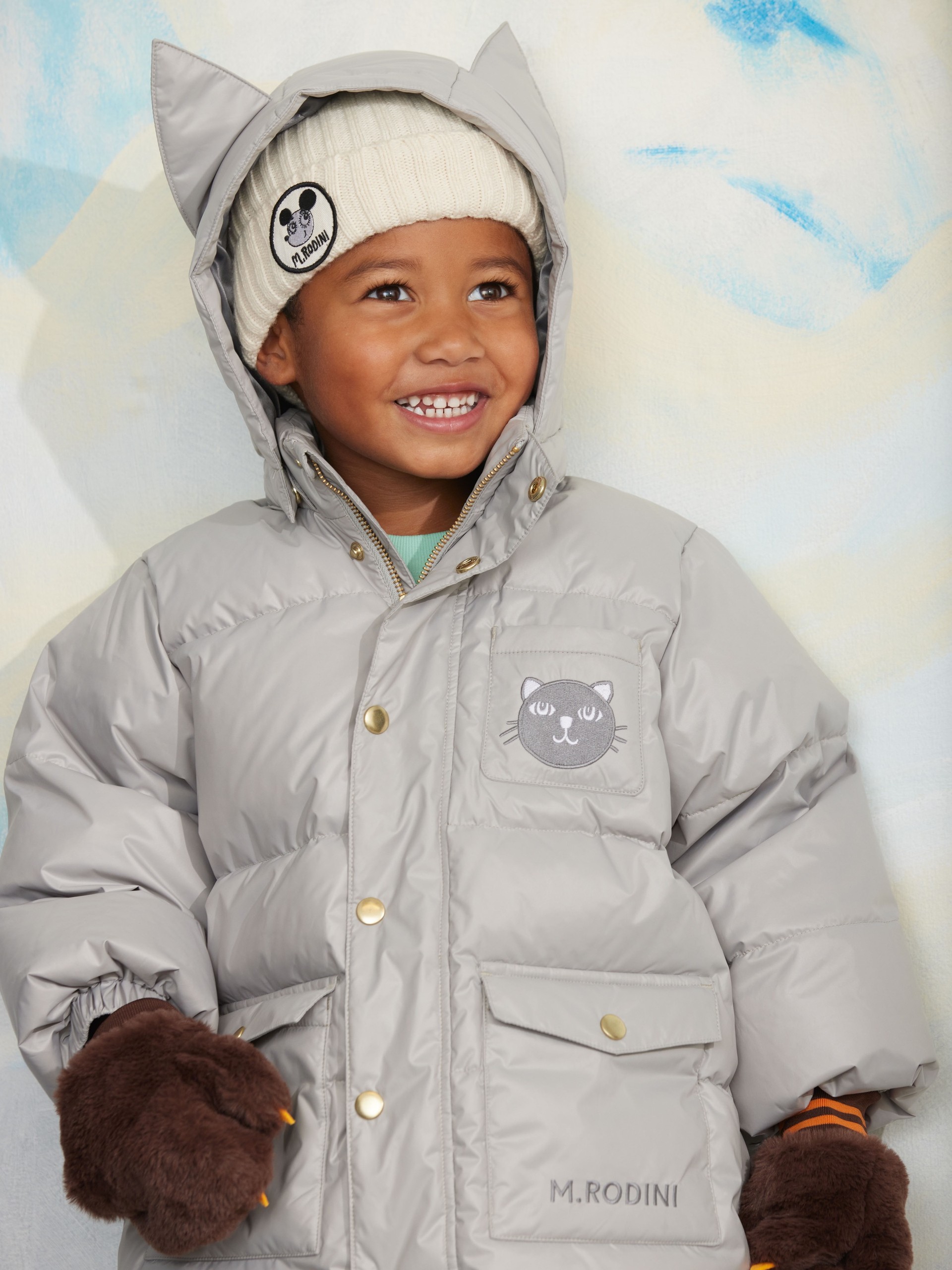 Child in a gray puffer from Mini Rodini with cat ears, wearing a white beanie and brown mittens, smiles against a soft blue and yellow background.