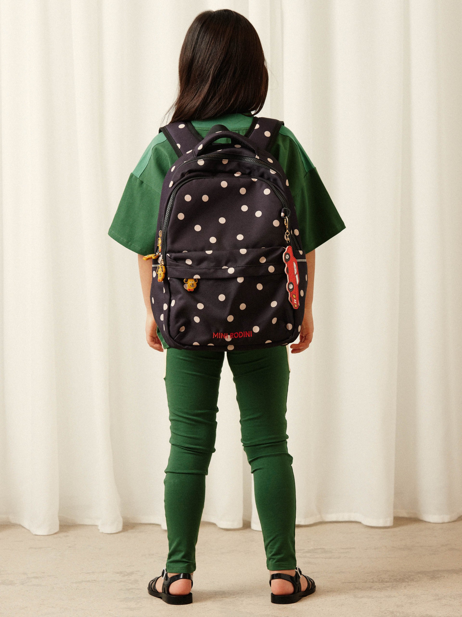 Child facing away, wearing a green outfit with a black polka dot backpack, standing on a beige carpet against a white curtain backdrop.