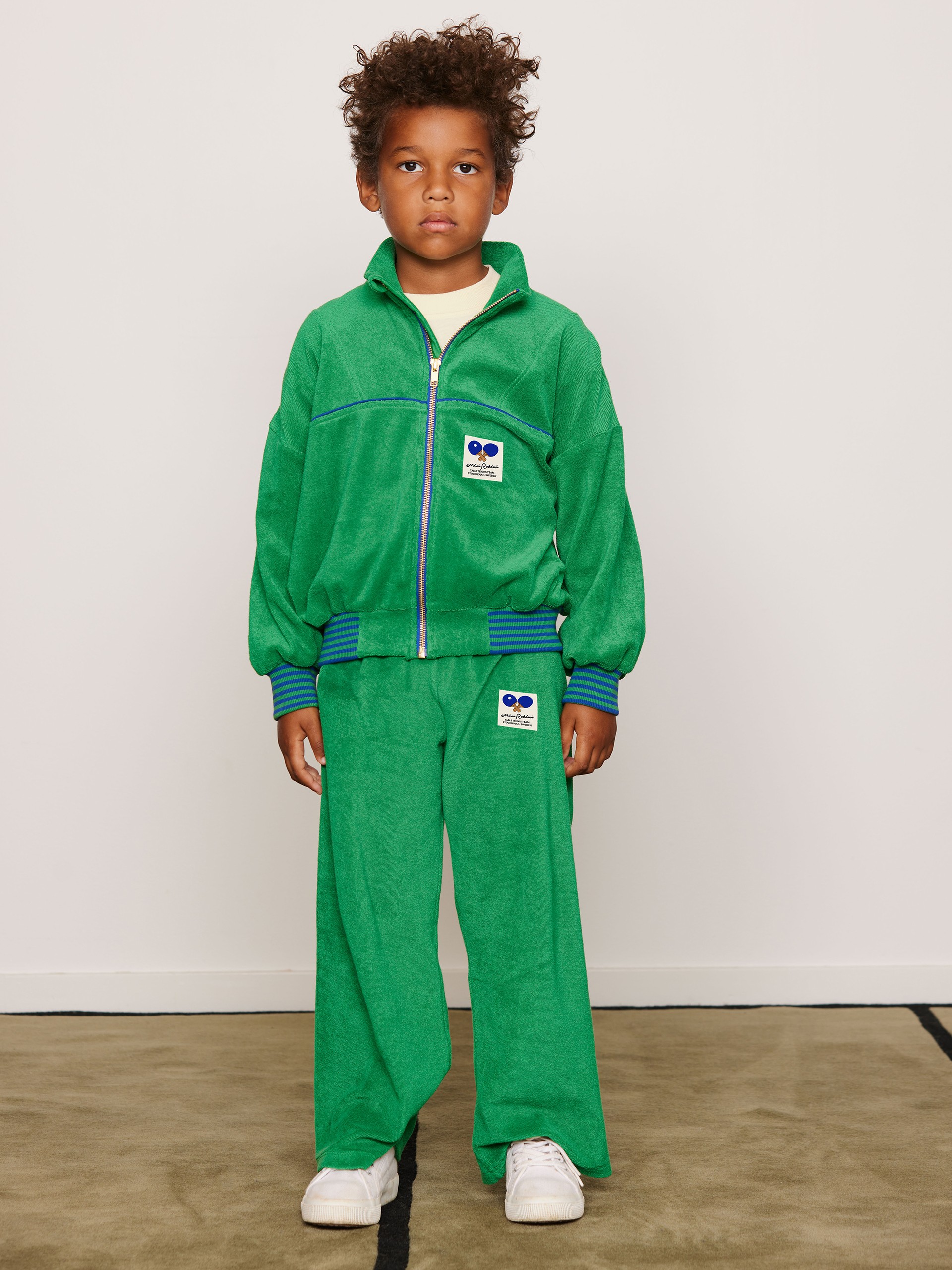 Young child wearing a bright green Terry tracksuit from Mini Rodini Table Tennis collectionwith, standing on a beige carpet, white background.