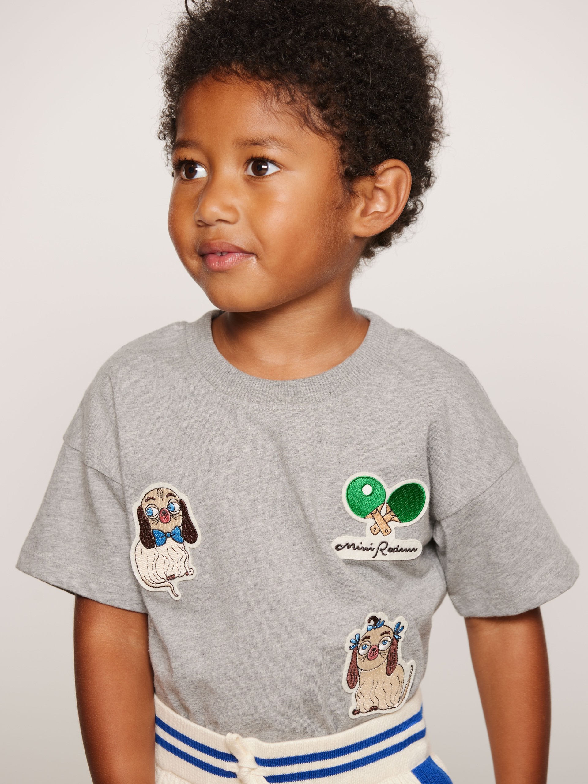 Child wearing a grey T-shirt with Mini Rodini dogs and ping pong paddle patches, looking to the side.