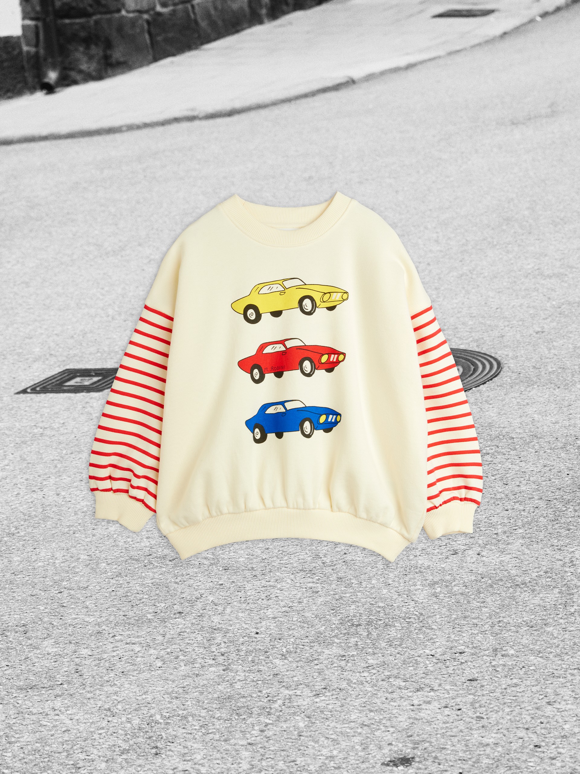 Cream sweater with red-striped sleeves featuring yellow, red, and blue car prints, displayed on a street-like background.